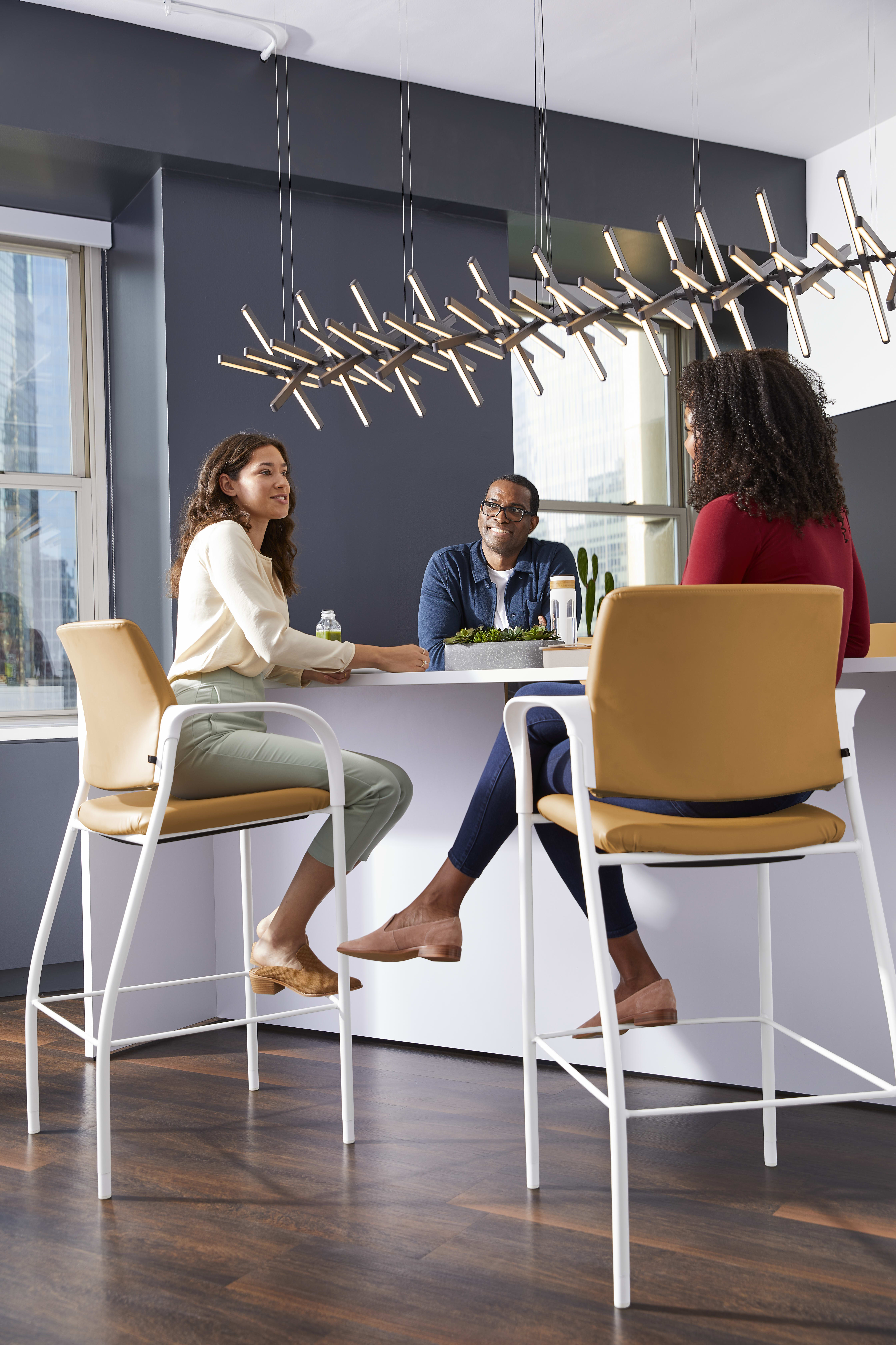 The image depicts a modern office space with a group of people seated at a table, engaged in conversation. The background features a unique lighting fixture and a dark-colored wall, creating a stylish and contemporary atmosphere.