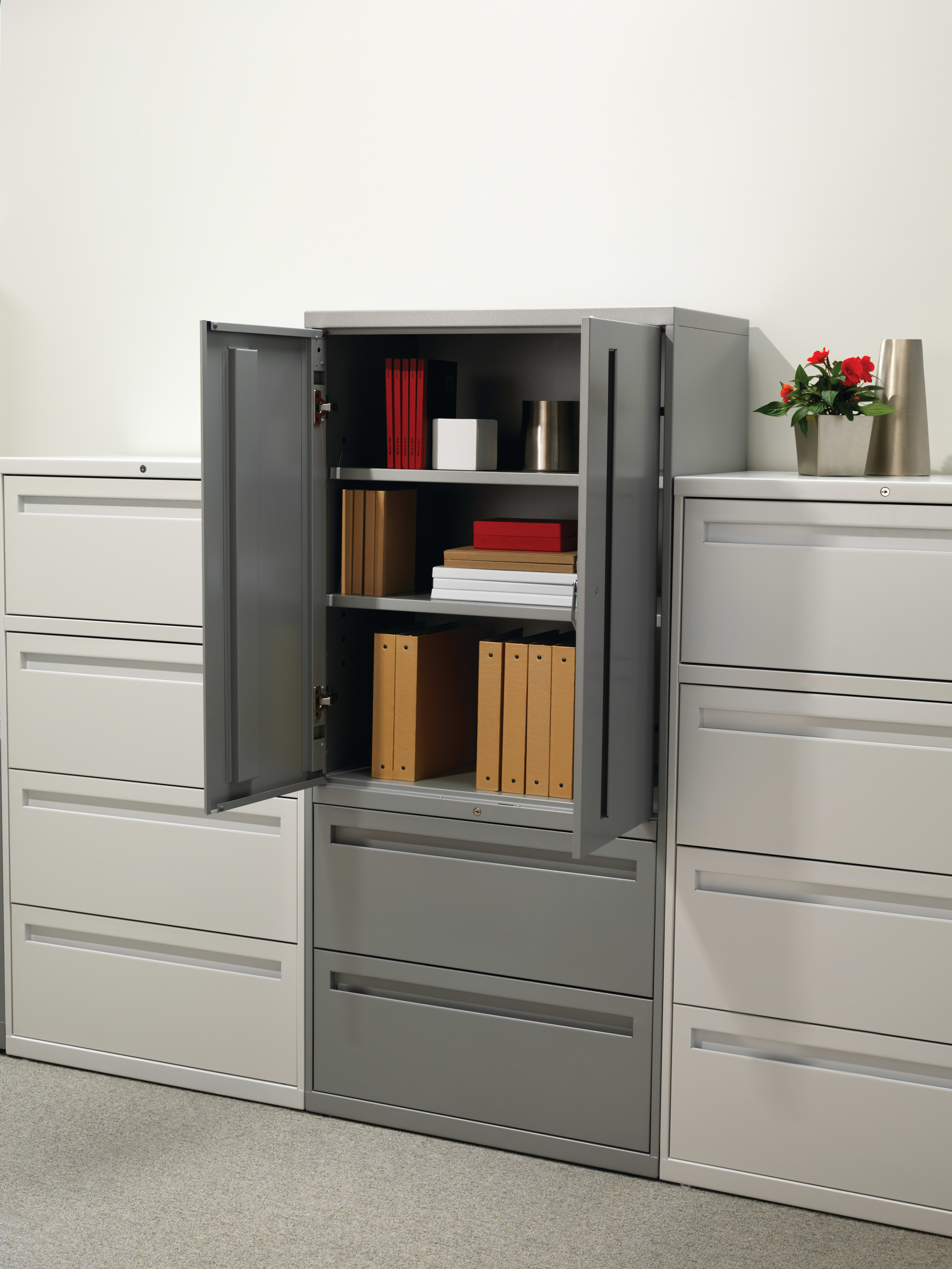 A gray metal storage cabinet with multiple drawers and shelves, containing various office supplies and documents, stands in a clean, minimalist office environment.