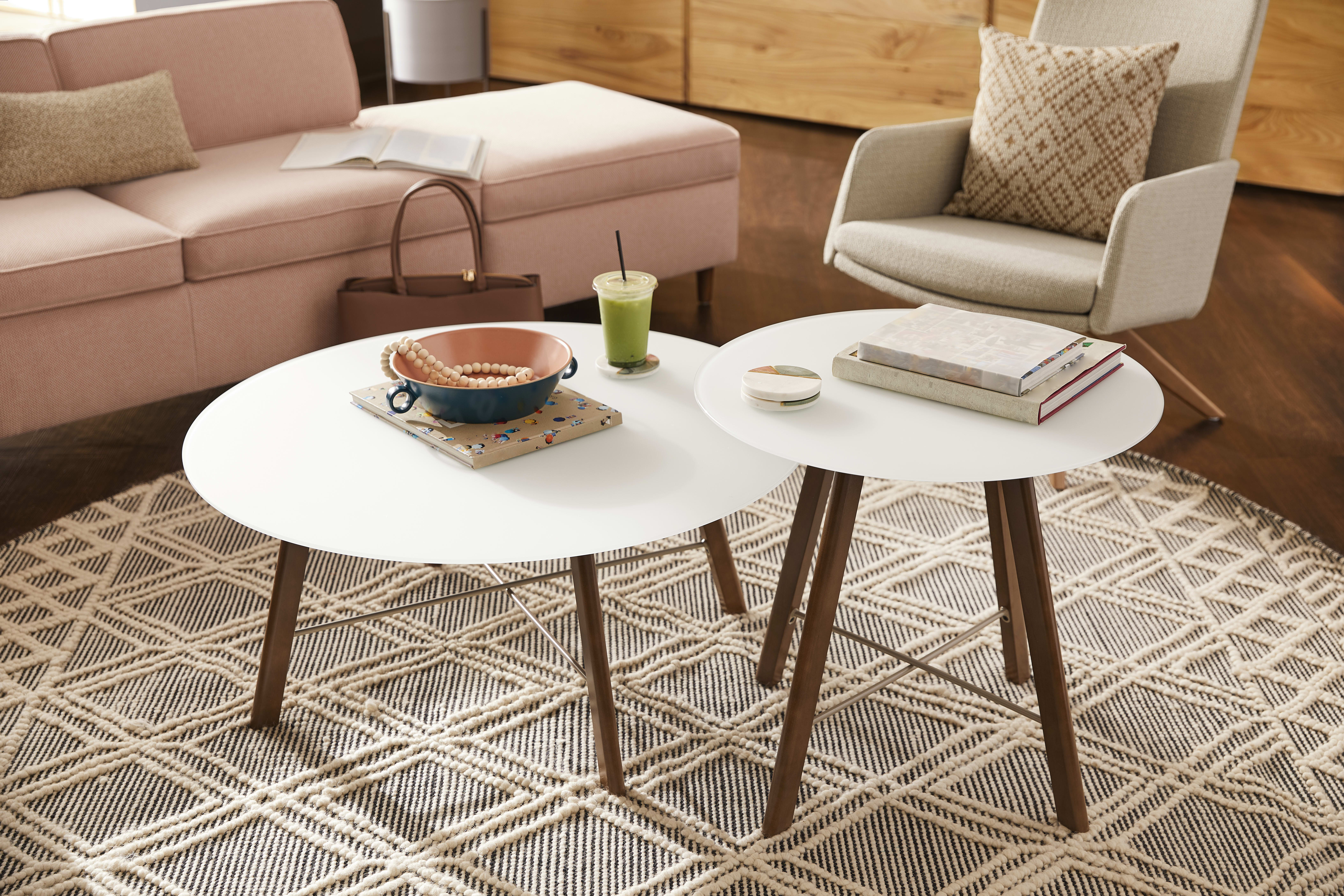 A cozy living room with a white round coffee table, a green smoothie, and a book on a patterned rug, surrounded by a beige sofa and a wooden cabinet in the background.