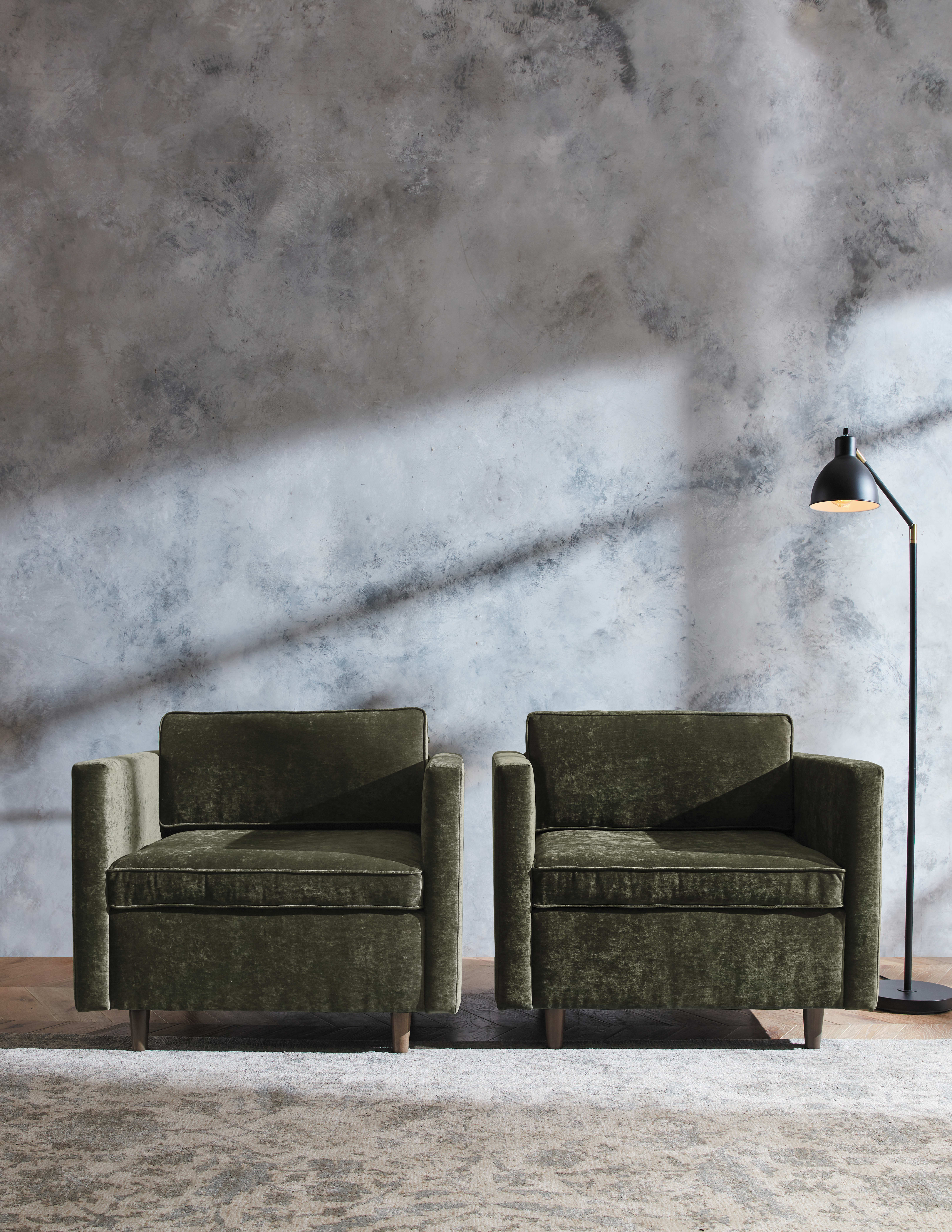 Two olive green leather armchairs sit in a minimalist room with a textured concrete wall in the background and a tall floor lamp illuminating the space.