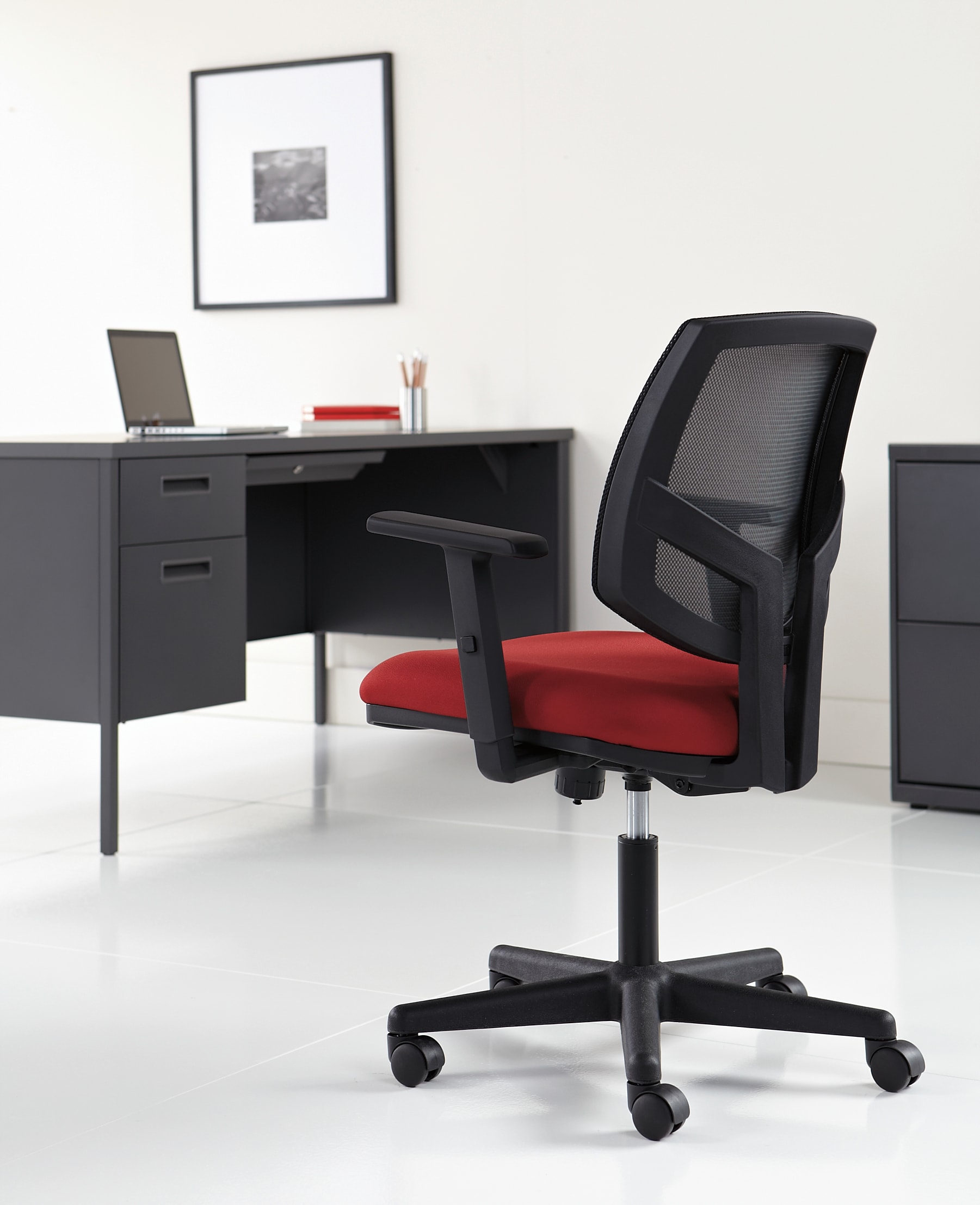 A modern, minimalist office setup with a black desk, a laptop, and a red and black office chair on a white floor, with a framed artwork on the wall in the background.