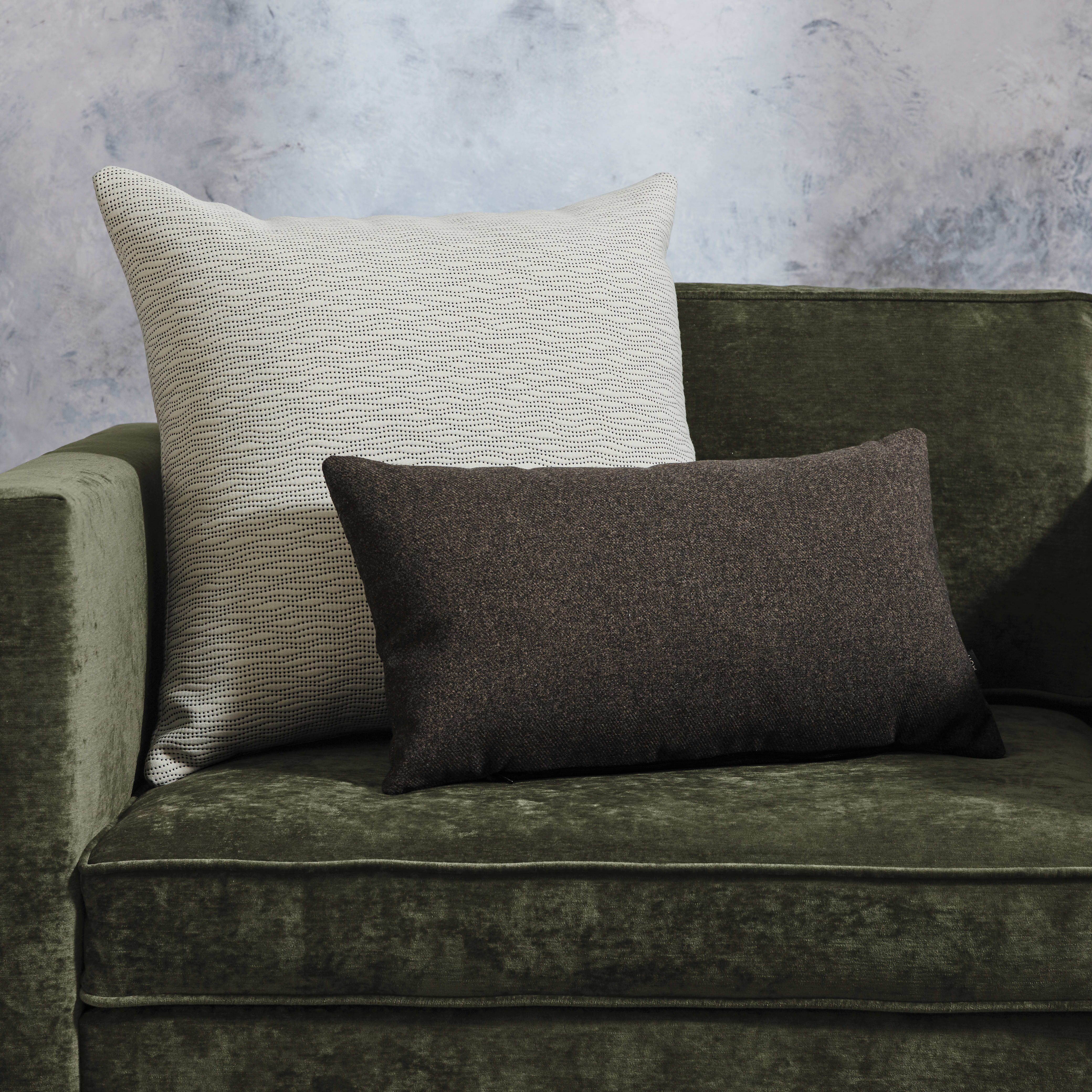 The image shows a green velvet sofa with two decorative pillows, one in a light beige pattern and the other in a dark brown solid color, against a plain gray background.