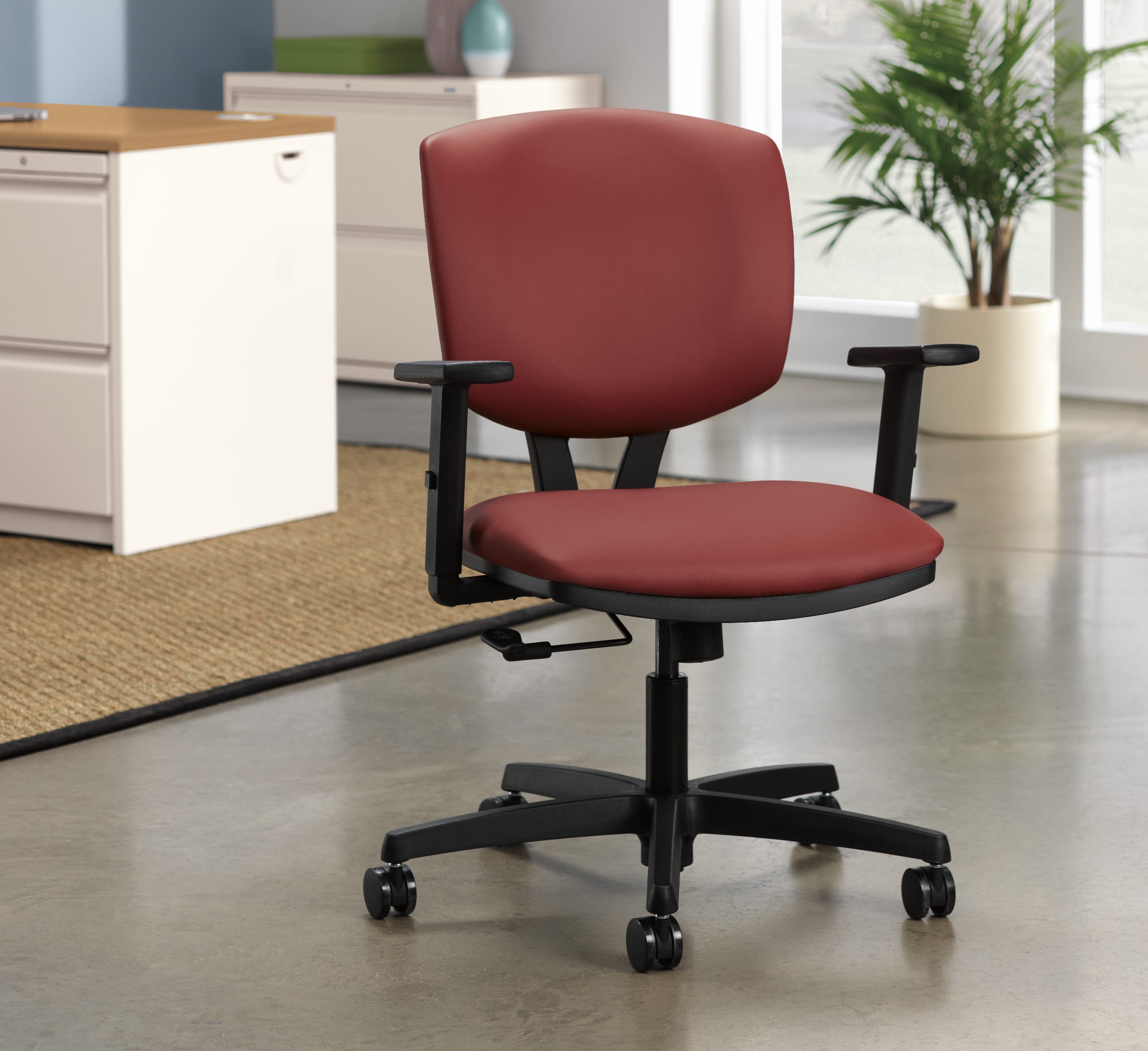 A red office chair with adjustable armrests sits on a tiled floor, with a filing cabinet and a potted plant visible in the background.