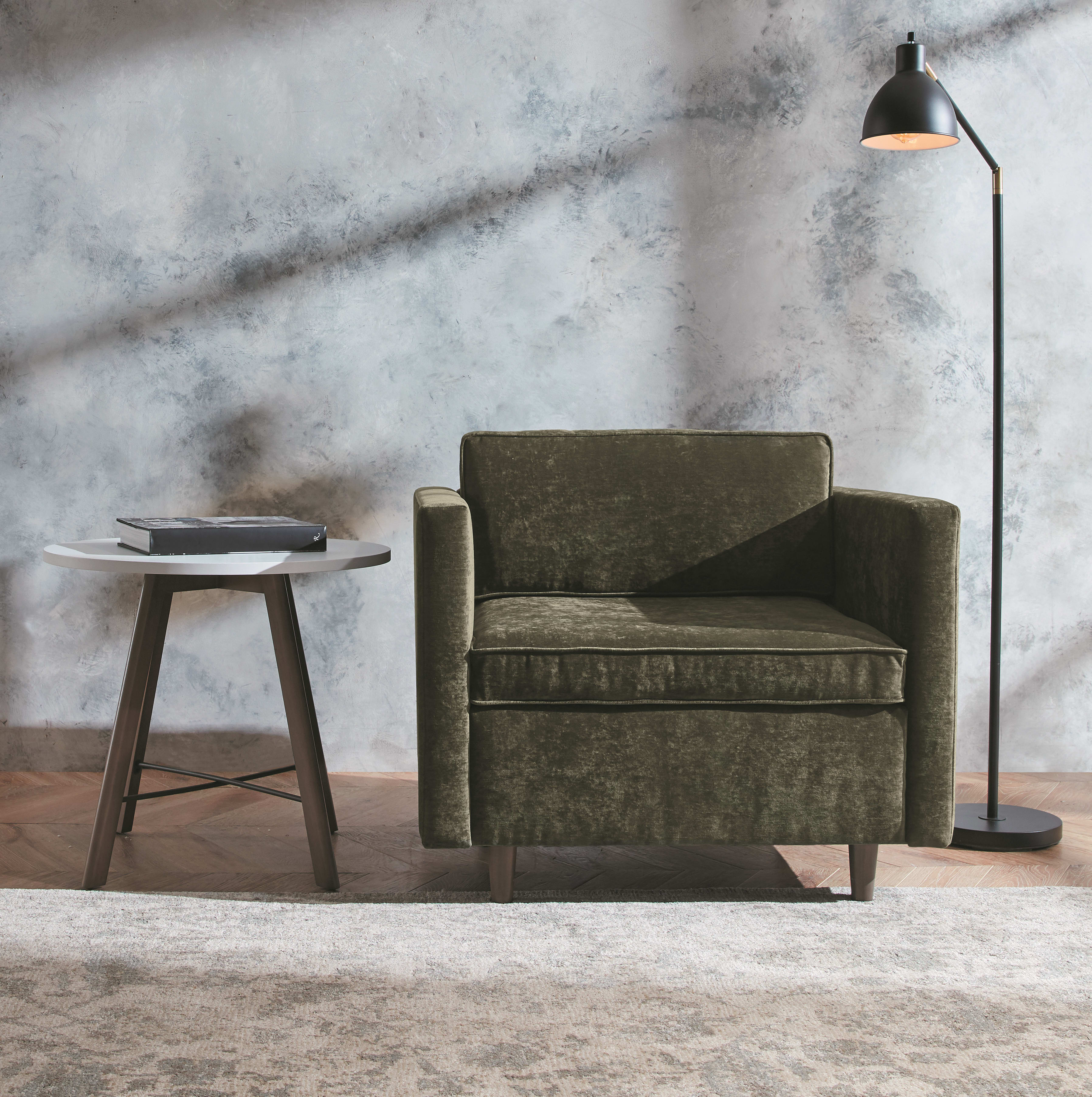A cozy living room setting with a green upholstered armchair, a round wooden side table, and a floor lamp against a textured marble-like wall backdrop.