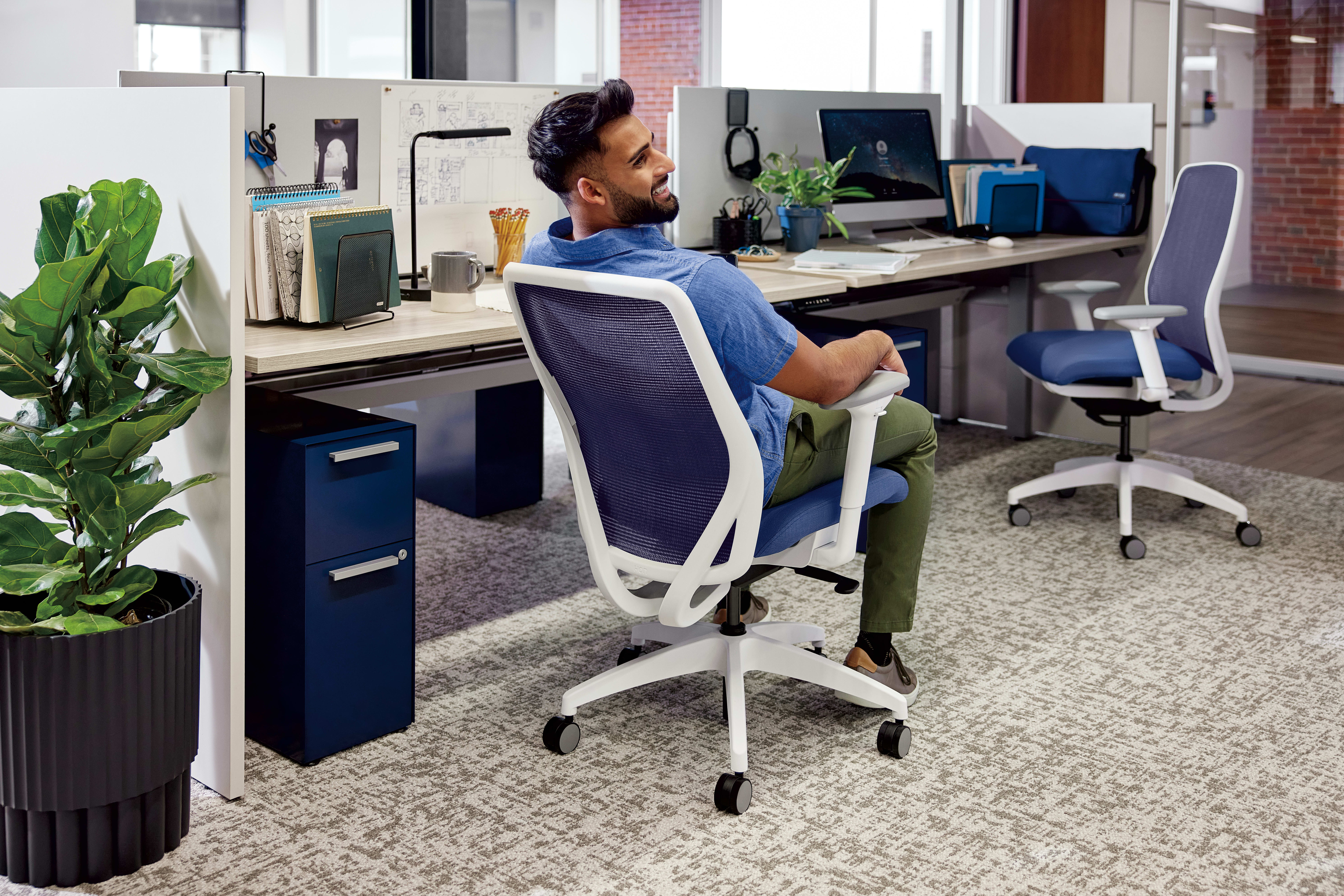 A person with a beard is sitting at a desk in an office environment, surrounded by various office equipment and a potted plant.