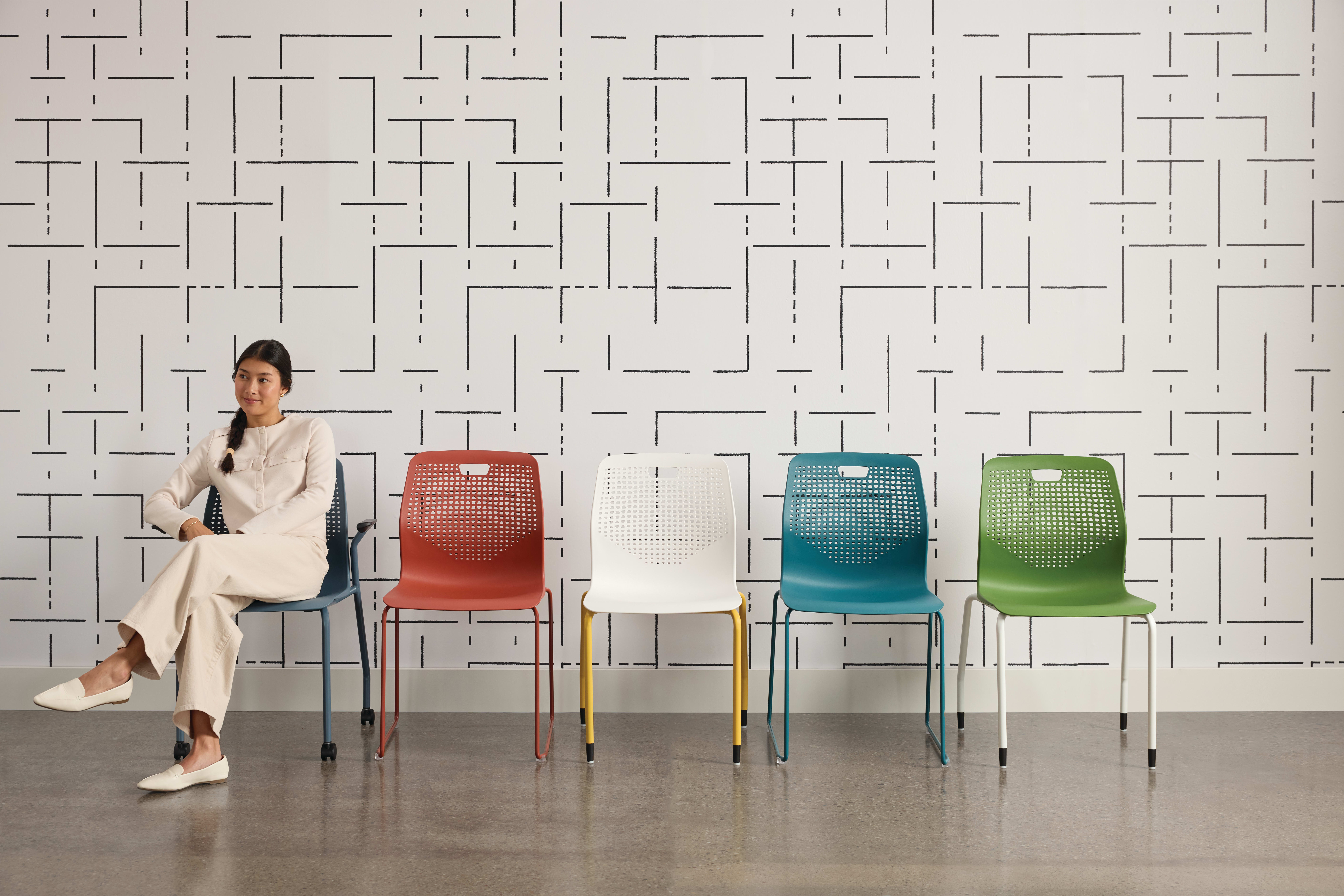 The image features a woman sitting on a chair in the foreground, surrounded by a patterned wall and a set of colorful chairs in the background.