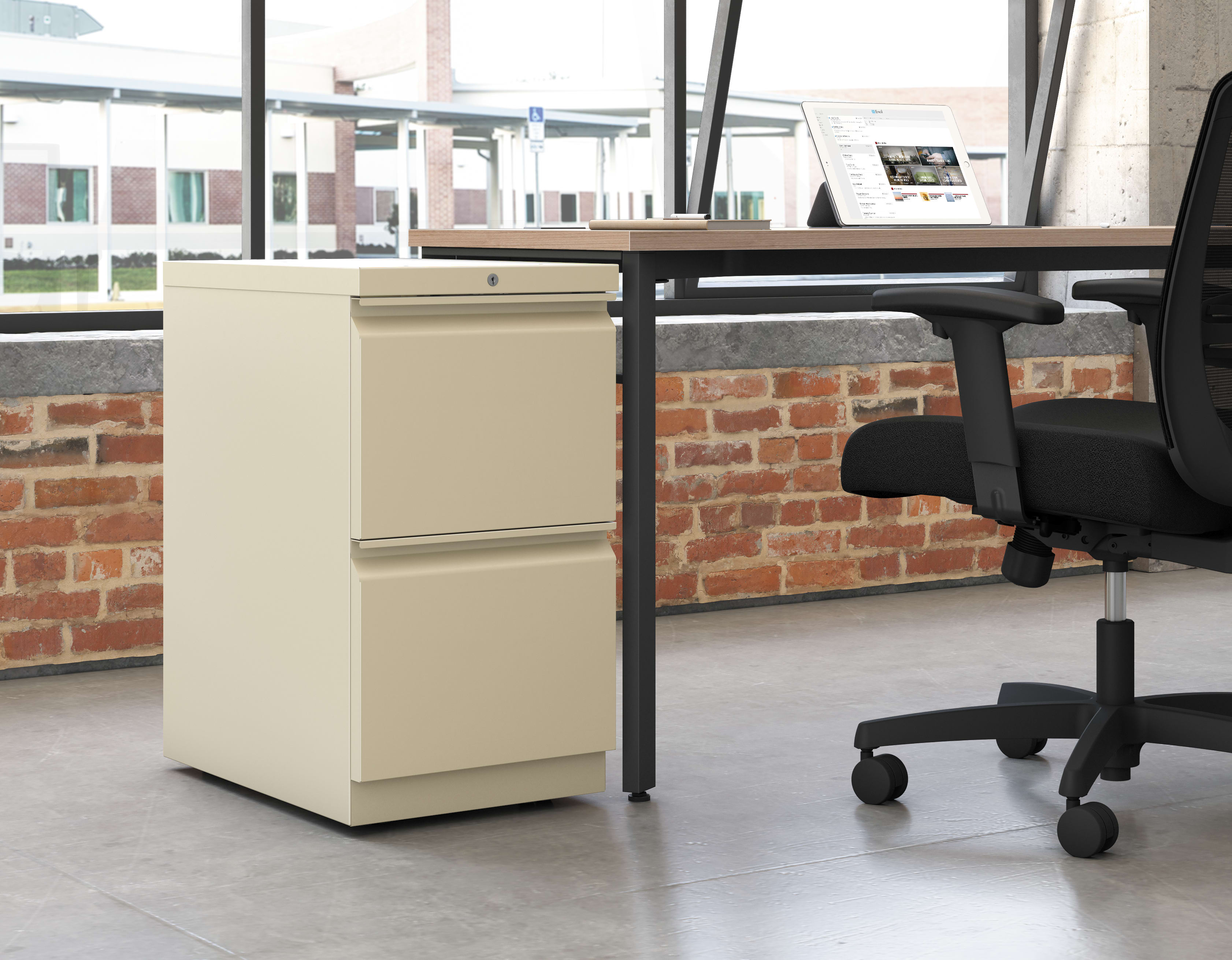 A modern office workspace with a wooden desk, a beige filing cabinet, and a black office chair set against a backdrop of exposed brick walls and large windows.