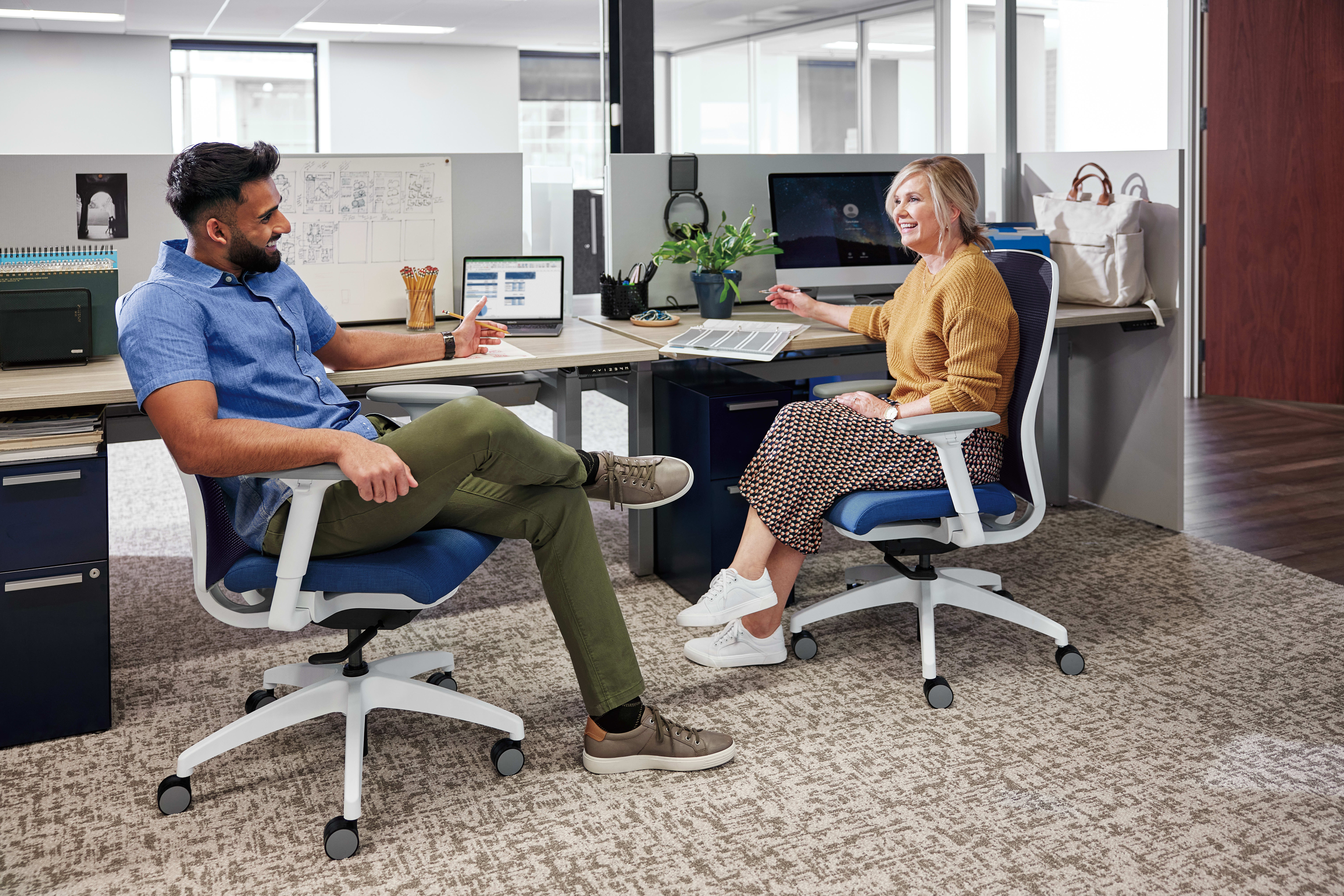 Two people, a man and a woman, are seated at desks in an office environment, engaged in conversation and working on computers.