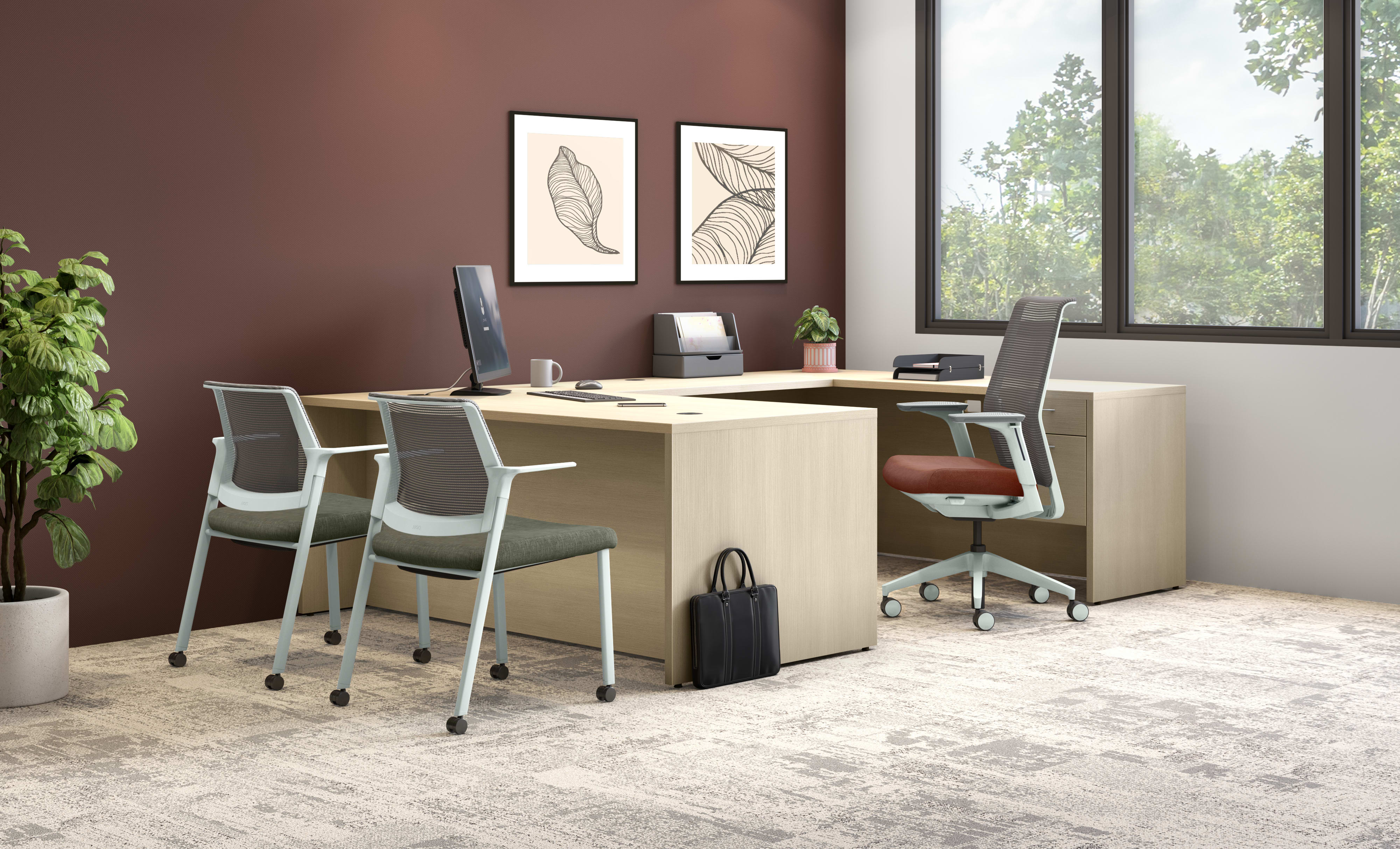 A modern and minimalist home office setup with a wooden desk, white chairs, and framed artwork on the burgundy-colored walls, set against a backdrop of large windows overlooking a lush, green outdoor environment.