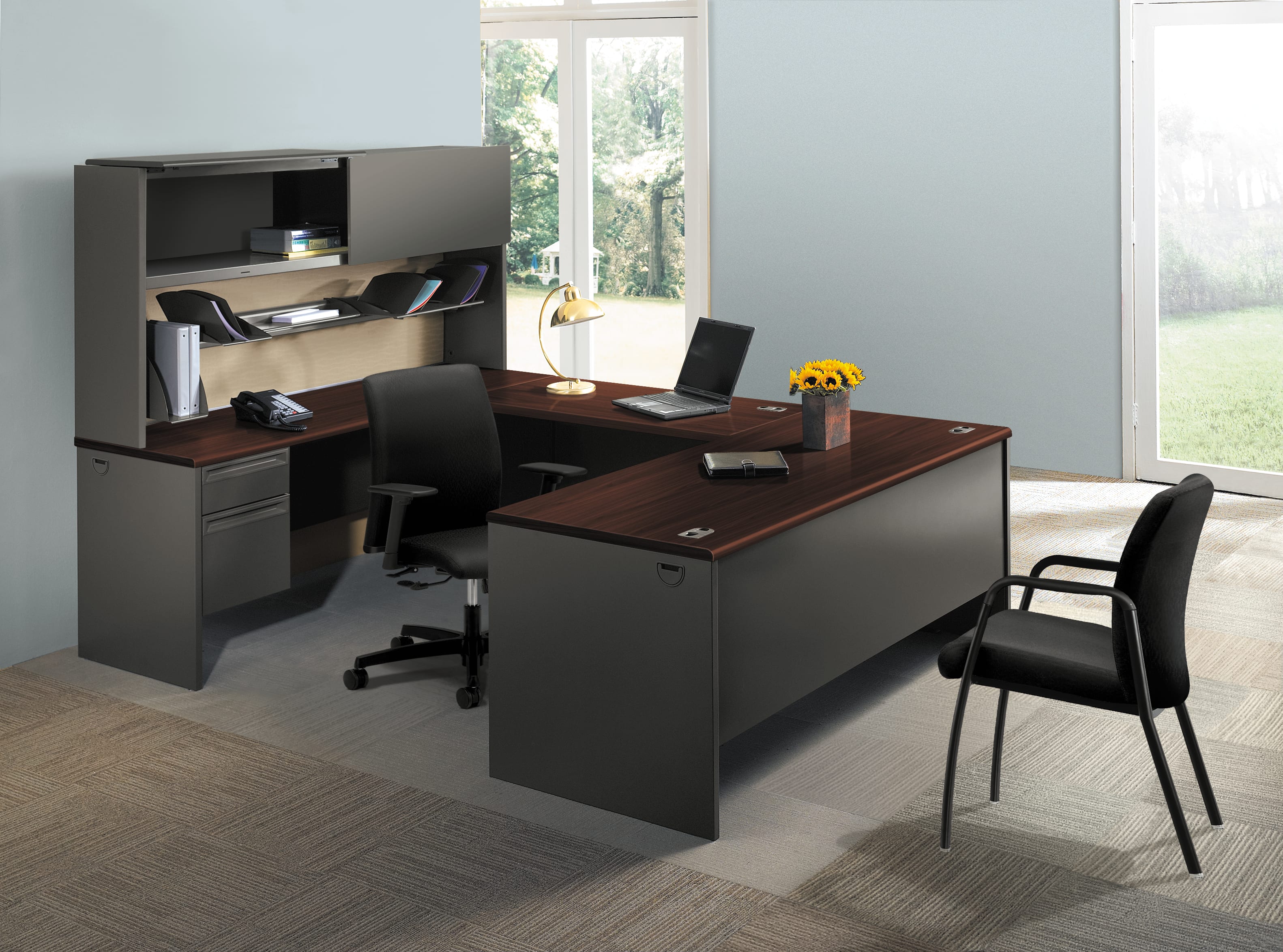 A modern and minimalist home office setup with a large wooden desk, a black computer chair, and a bookshelf unit against a bright, natural-lit background.