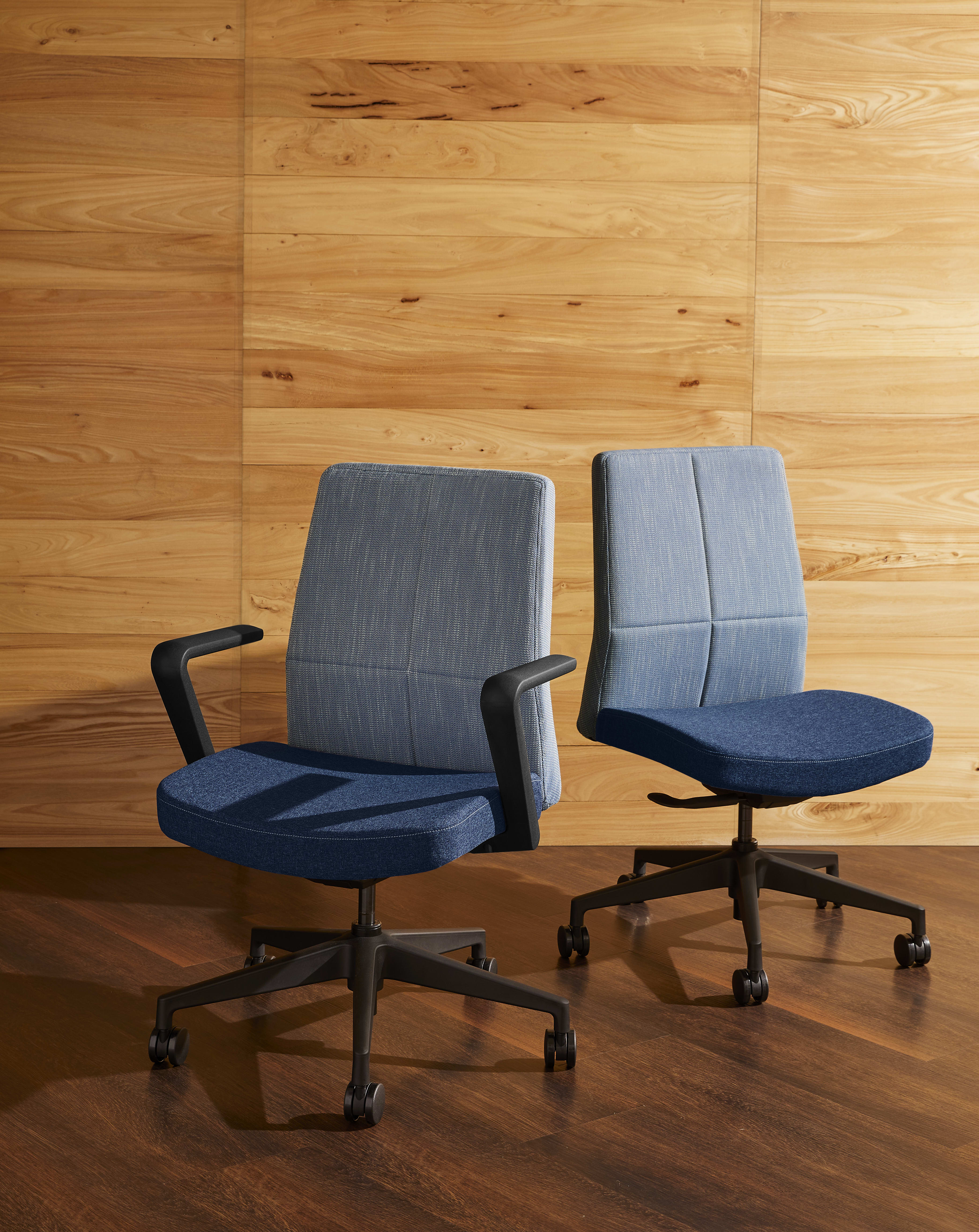 Two gray office chairs with armrests sit on a wooden floor against a wooden wall background.