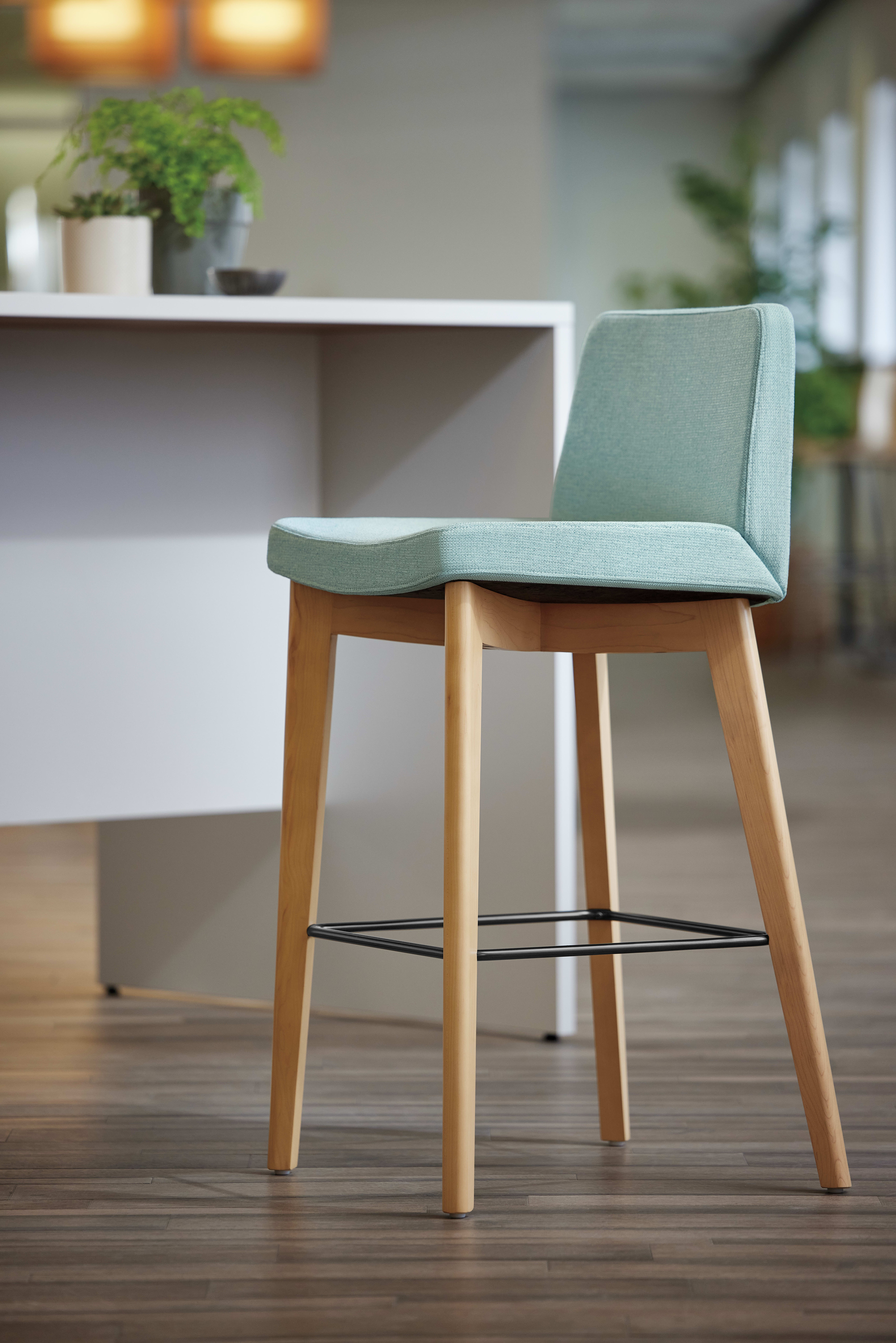 A modern, minimalist chair with a light green upholstered seat and wooden legs sits in a room with a white desk and potted plant in the background.