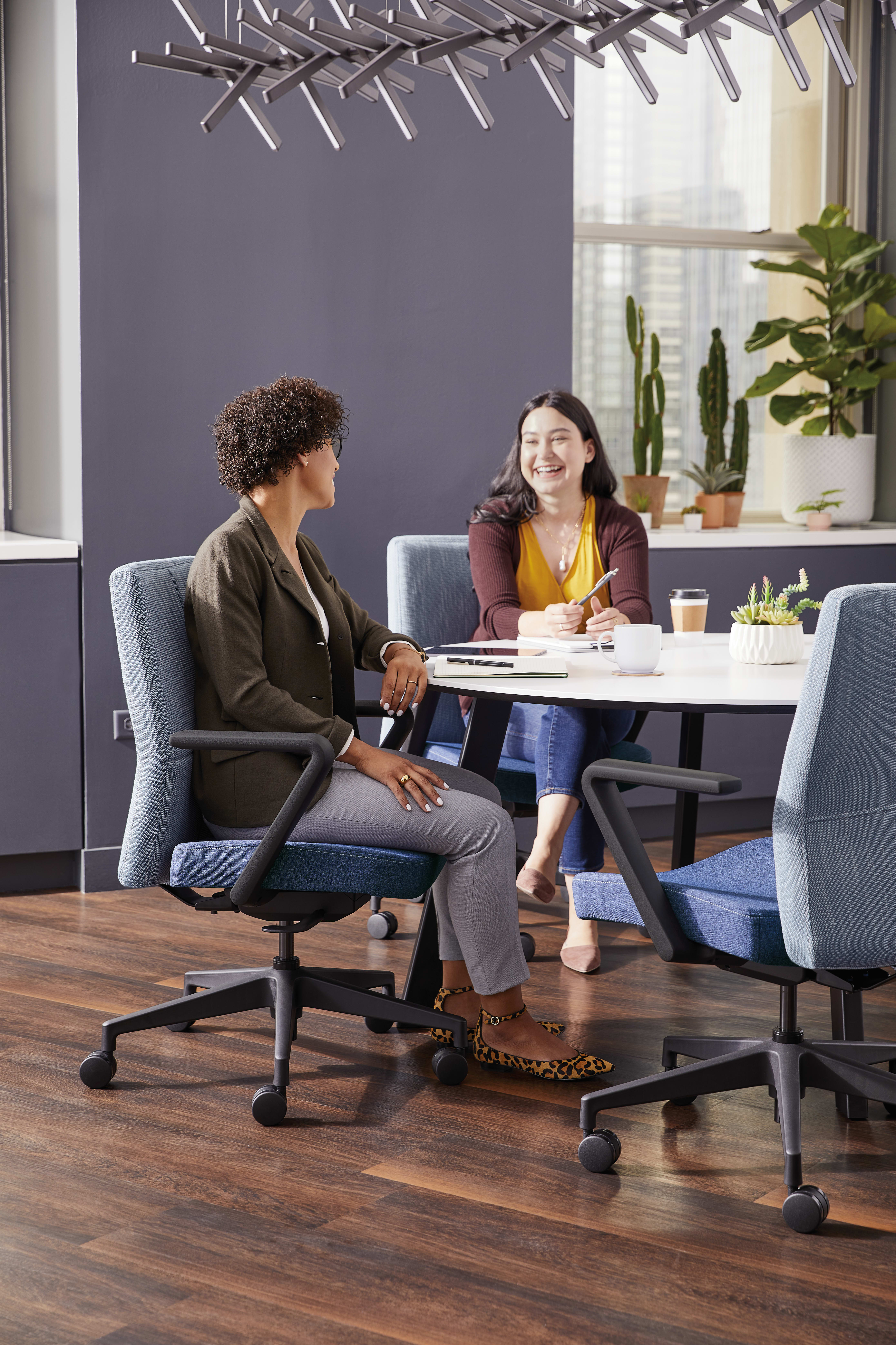 Two people, a woman and a man, are sitting at a table in an office-like setting, engaged in conversation. The background features a gray wall, potted plants, and a modern lighting fixture.