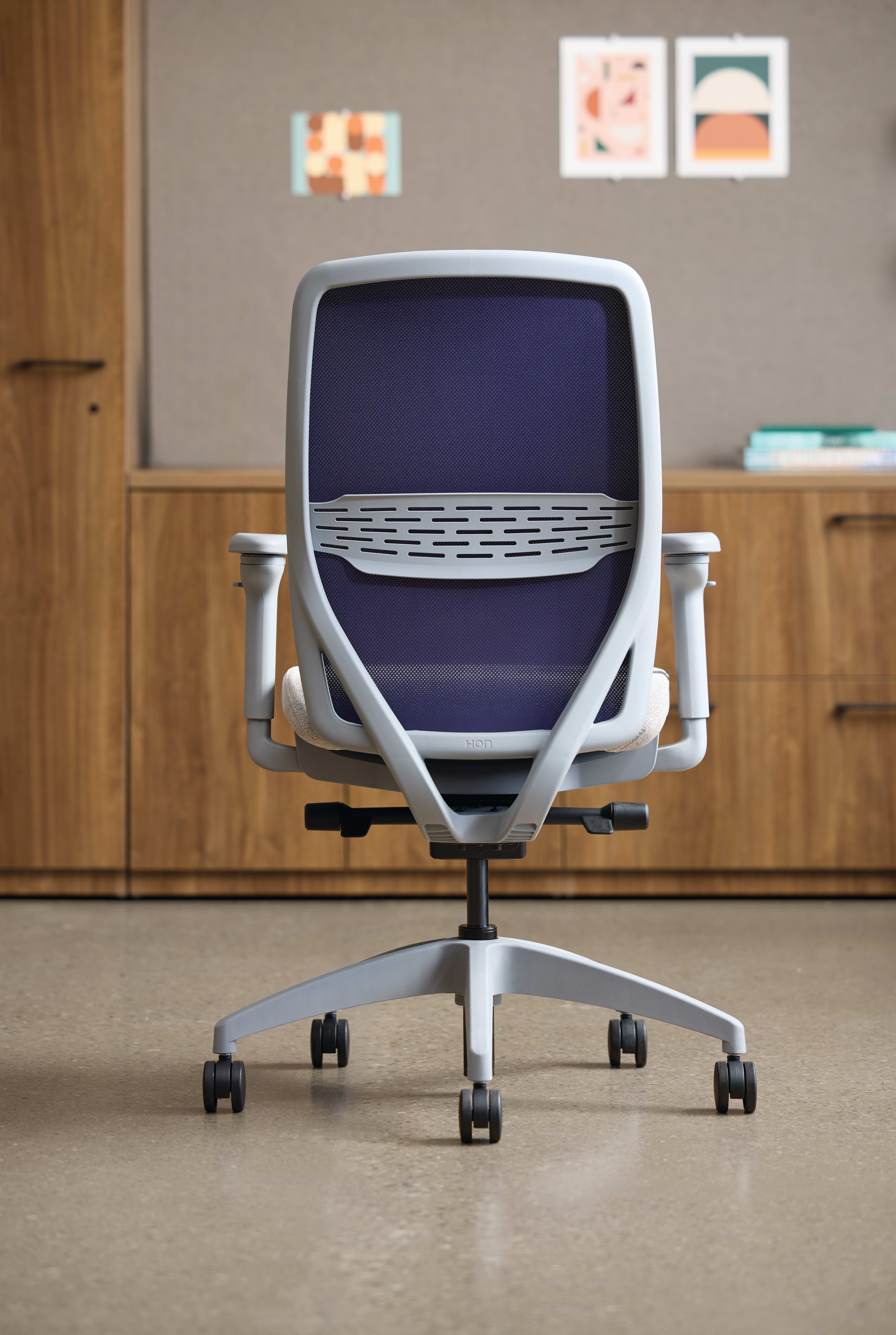 A modern, ergonomic office chair with a purple mesh backrest and seat, situated in a minimalist workspace with wooden cabinets and framed artwork on the walls.