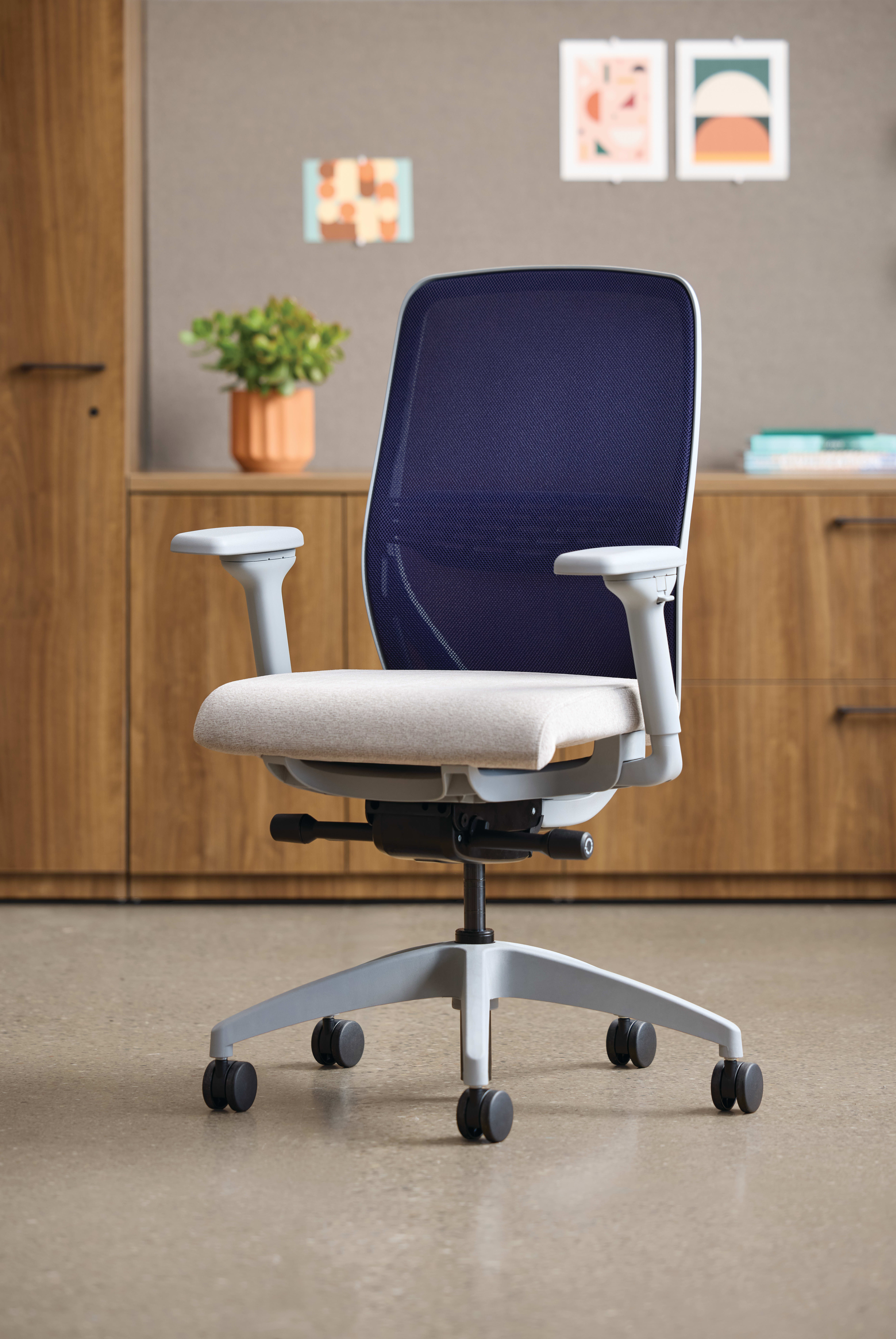 A modern, adjustable office chair with a navy blue backrest and a light-colored seat sits in a cozy, well-furnished room with wooden cabinets and framed artwork on the walls.