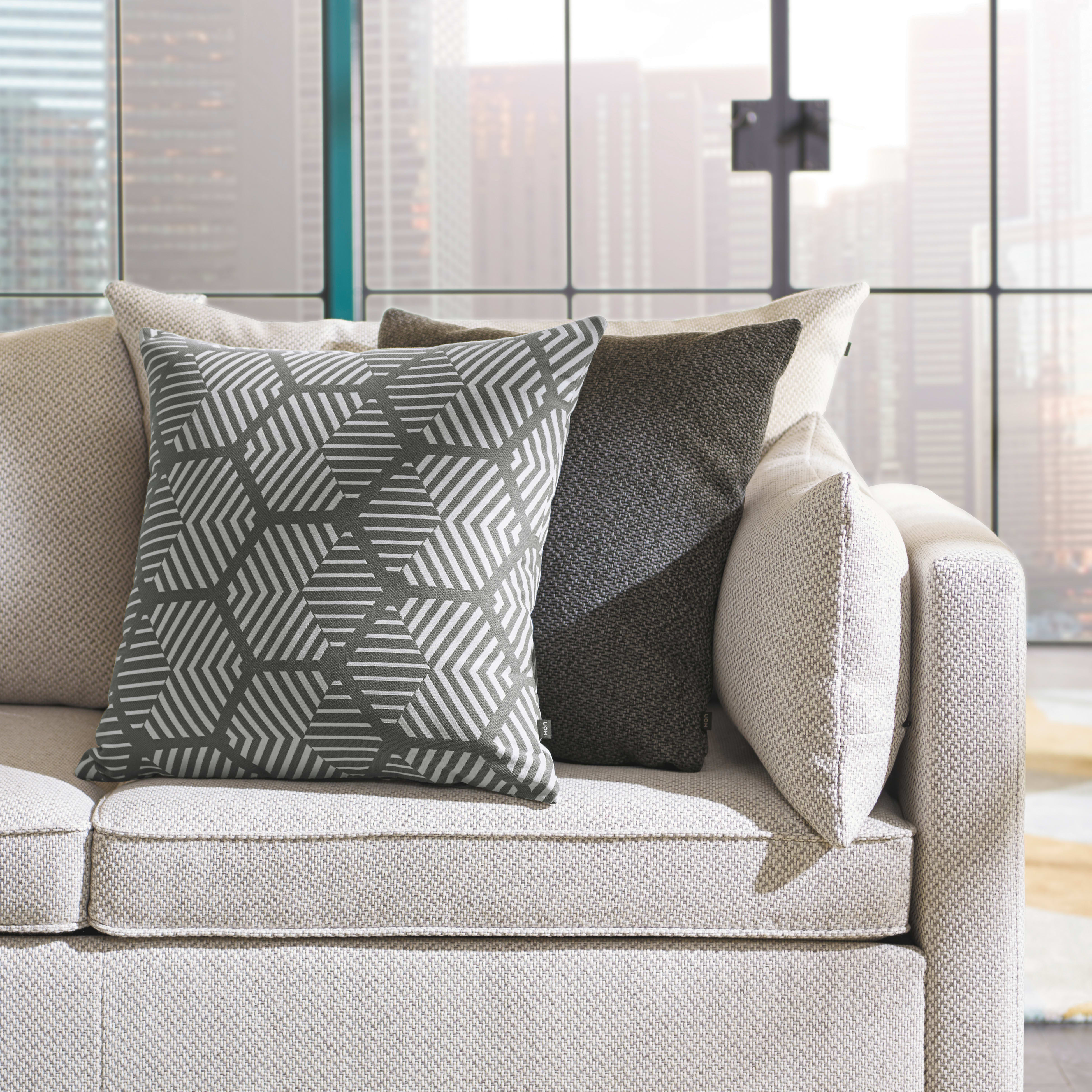 A cozy living room with a beige sofa adorned with patterned and textured throw pillows, set against a backdrop of large windows overlooking a cityscape.