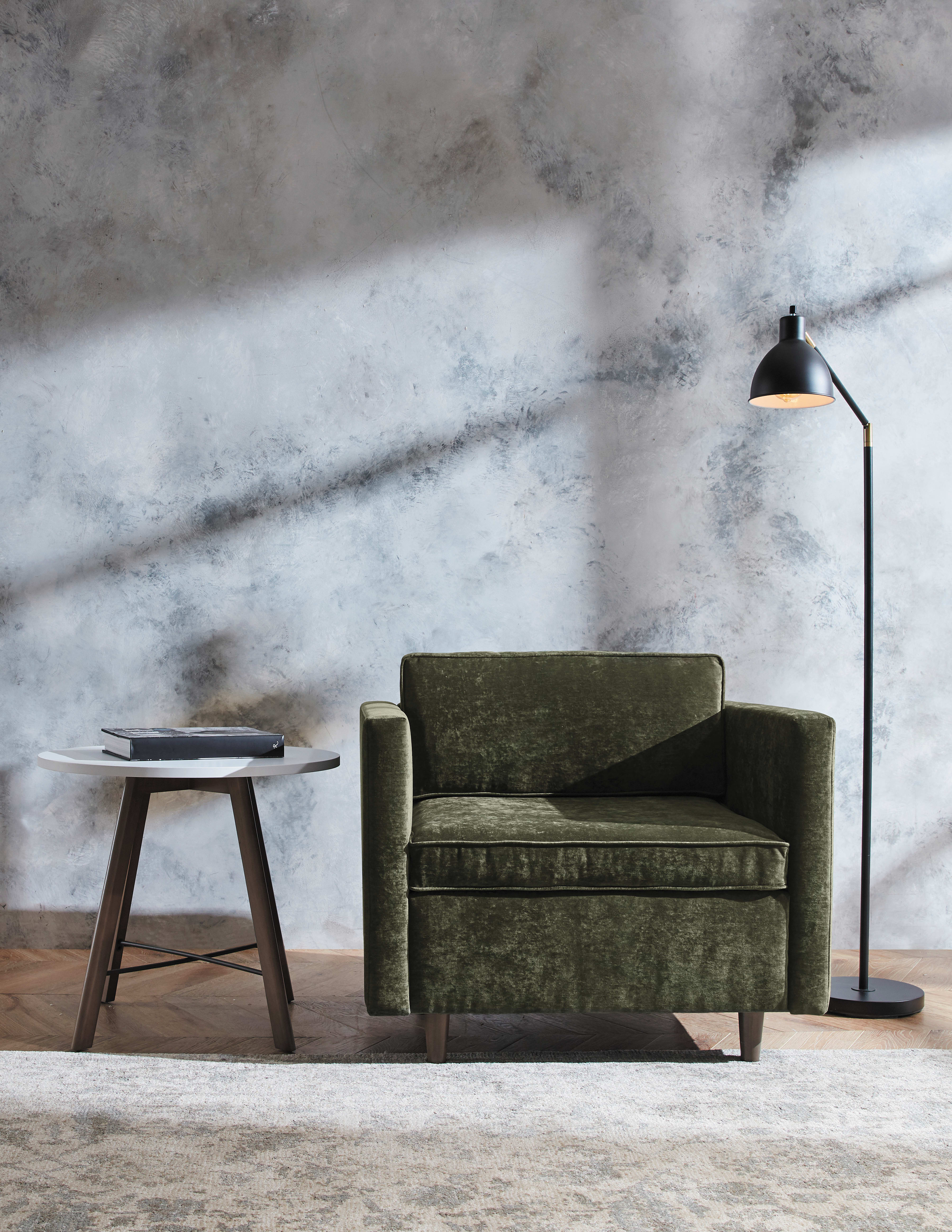 A cozy green velvet sofa sits in a dimly lit room, with a small wooden side table and a floor lamp providing a warm glow against a textured, gray wall backdrop.