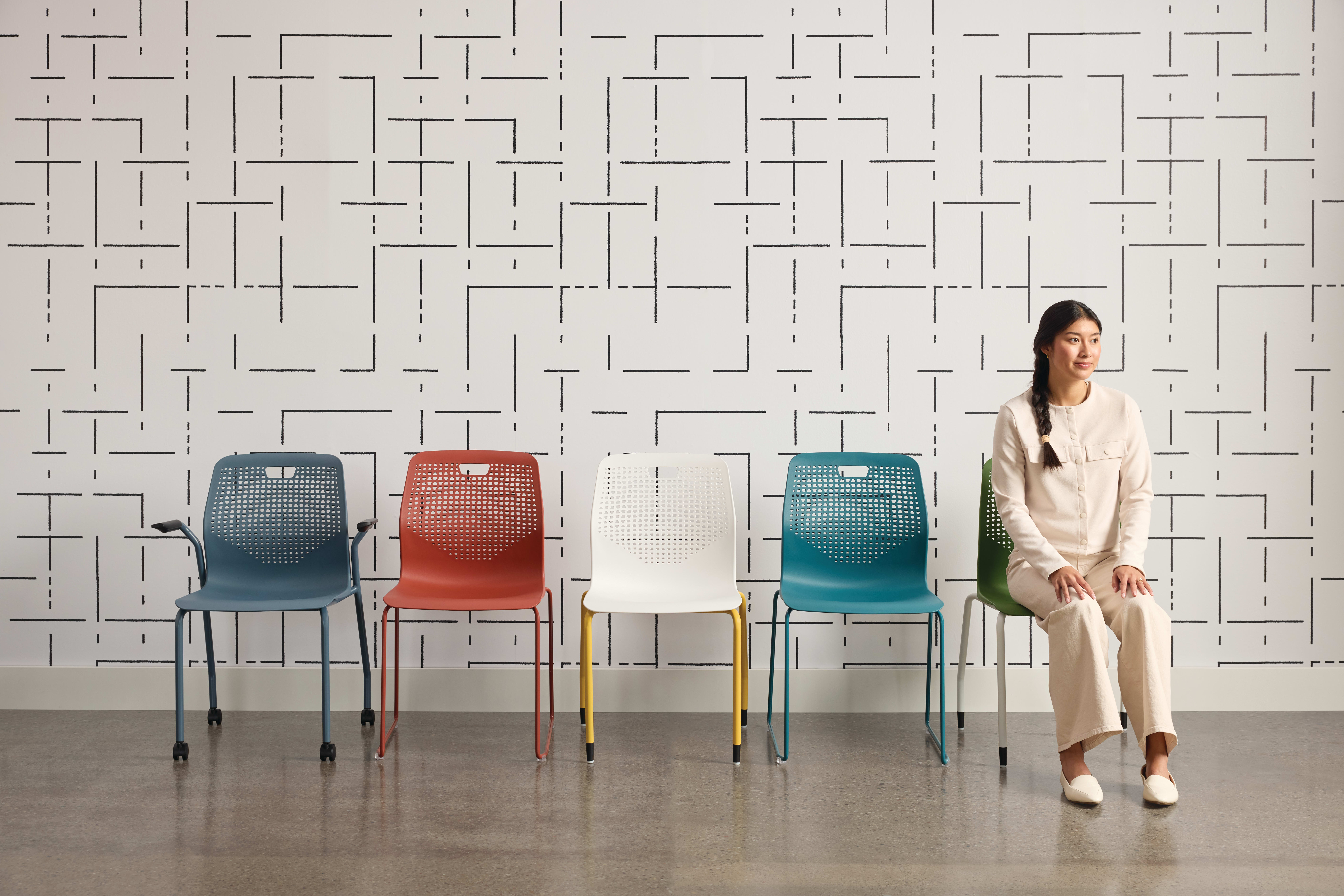 The image features a woman standing in front of a patterned wall, with four colorful chairs arranged in the foreground.