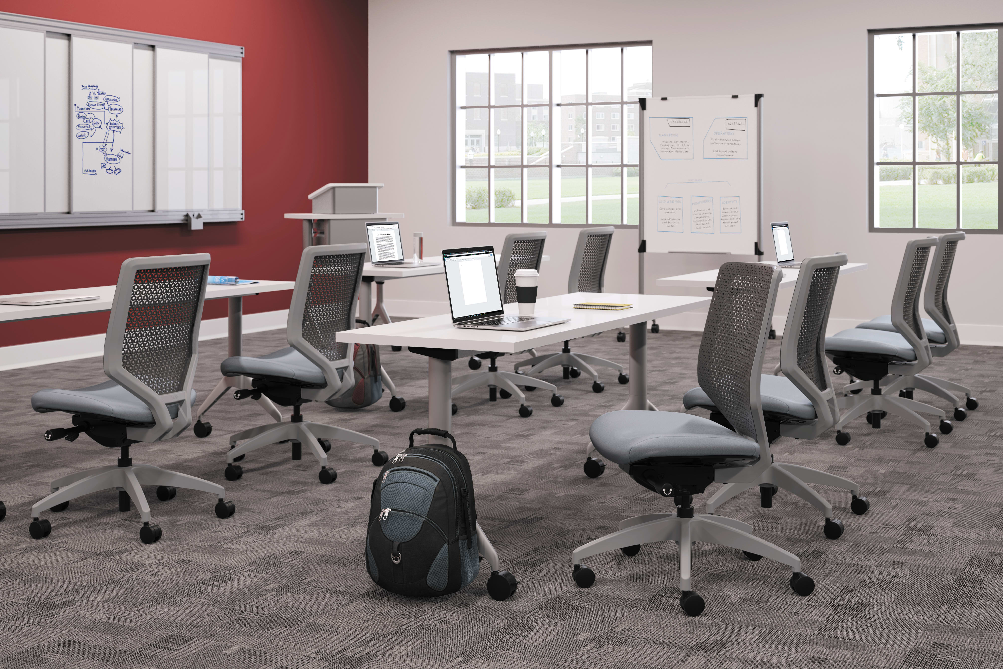 A modern, well-equipped office space with desks, chairs, and various office equipment, set against a red wall and large windows providing natural light.