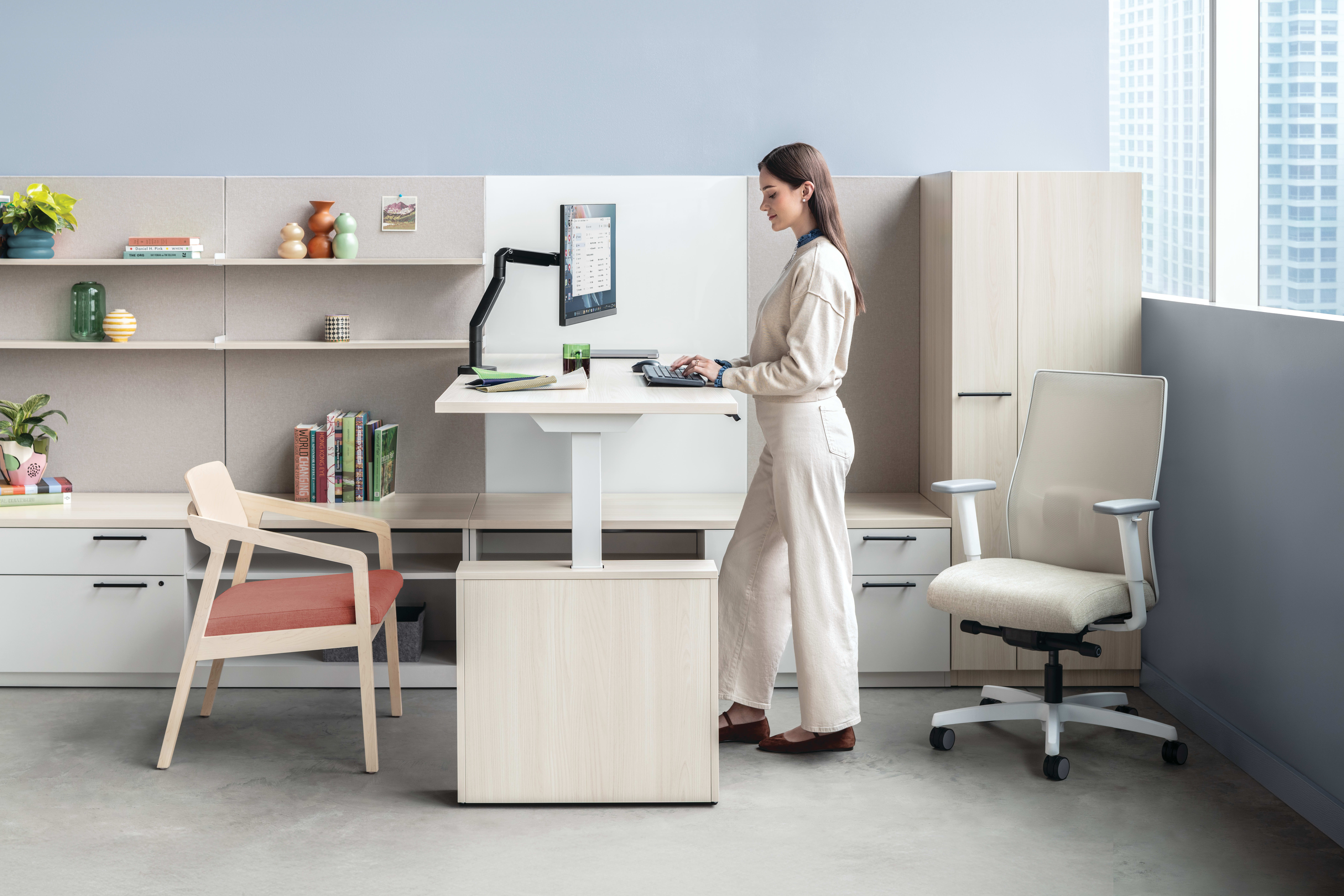 The image depicts a modern, minimalist home office setup with a woman standing at a standing desk, surrounded by shelves, chairs, and other office equipment.