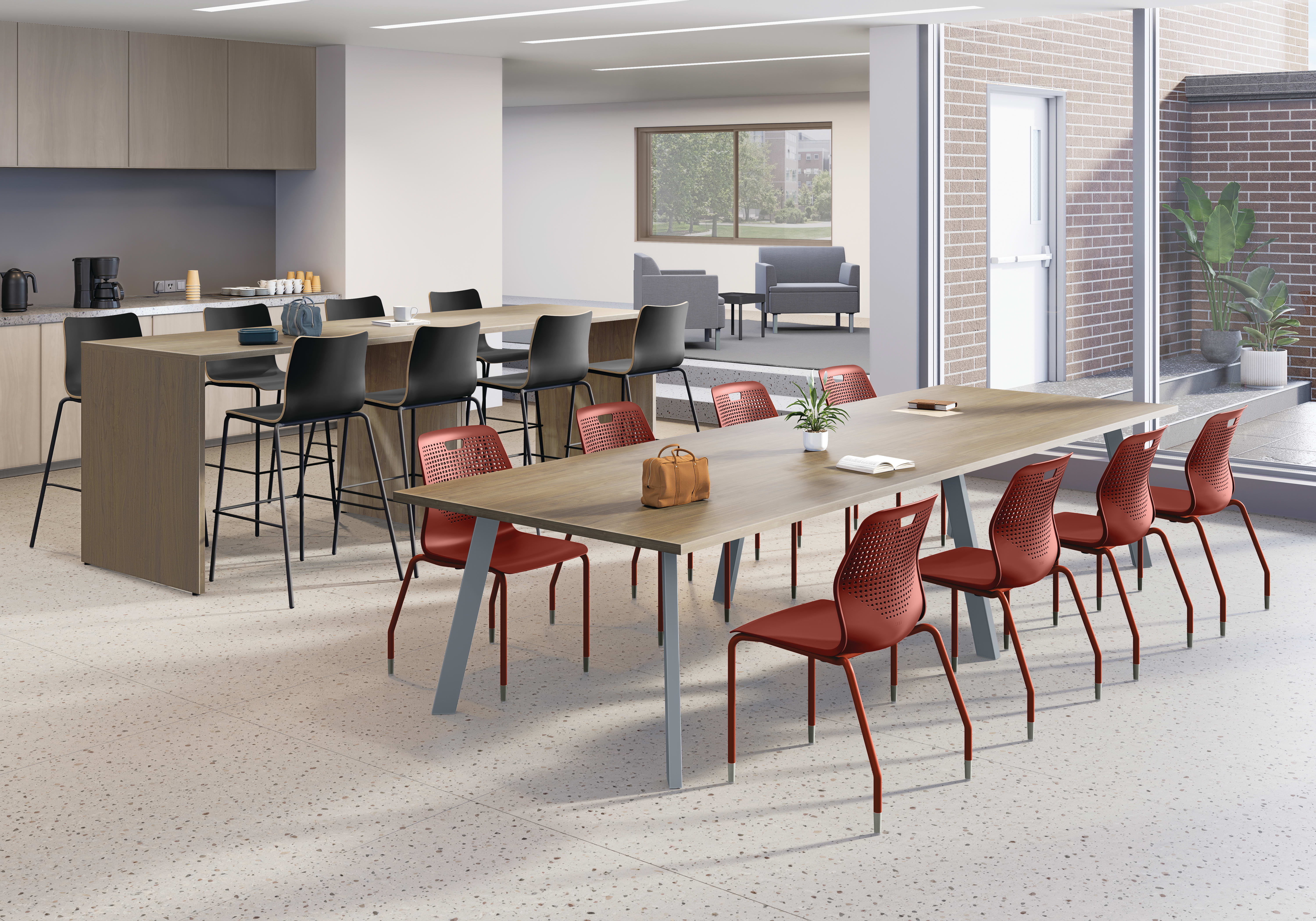 Modern collaborative workspace with long wooden tables, red and black chairs, coffee station, adjacent lounge seating, large windows, and bright contemporary lighting.