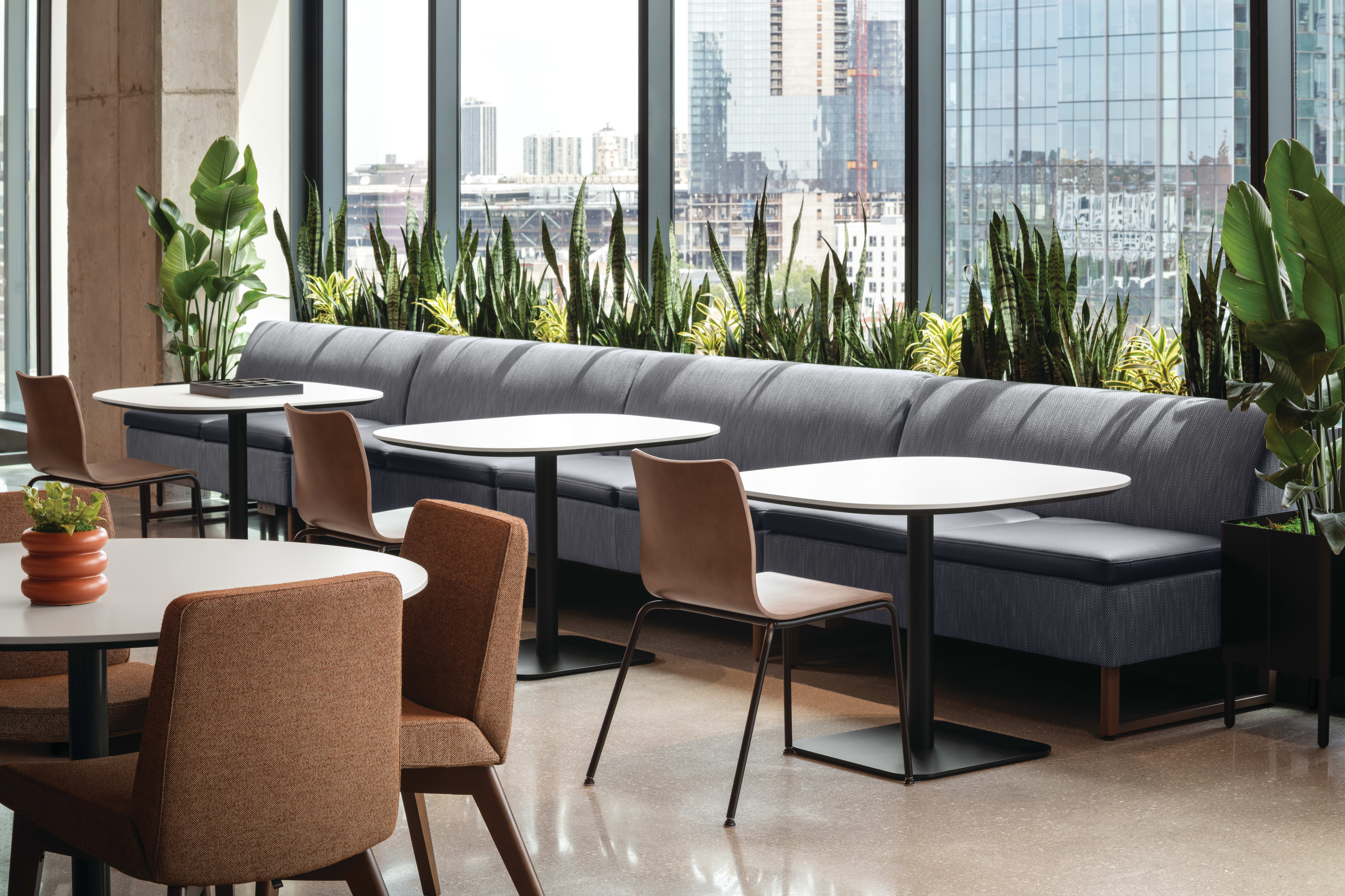 Café-style seating area with upholstered banquette, round pedestal tables, and mixed-material guest chairs, complemented by integrated planter with greenery and adjacent lounge seating.