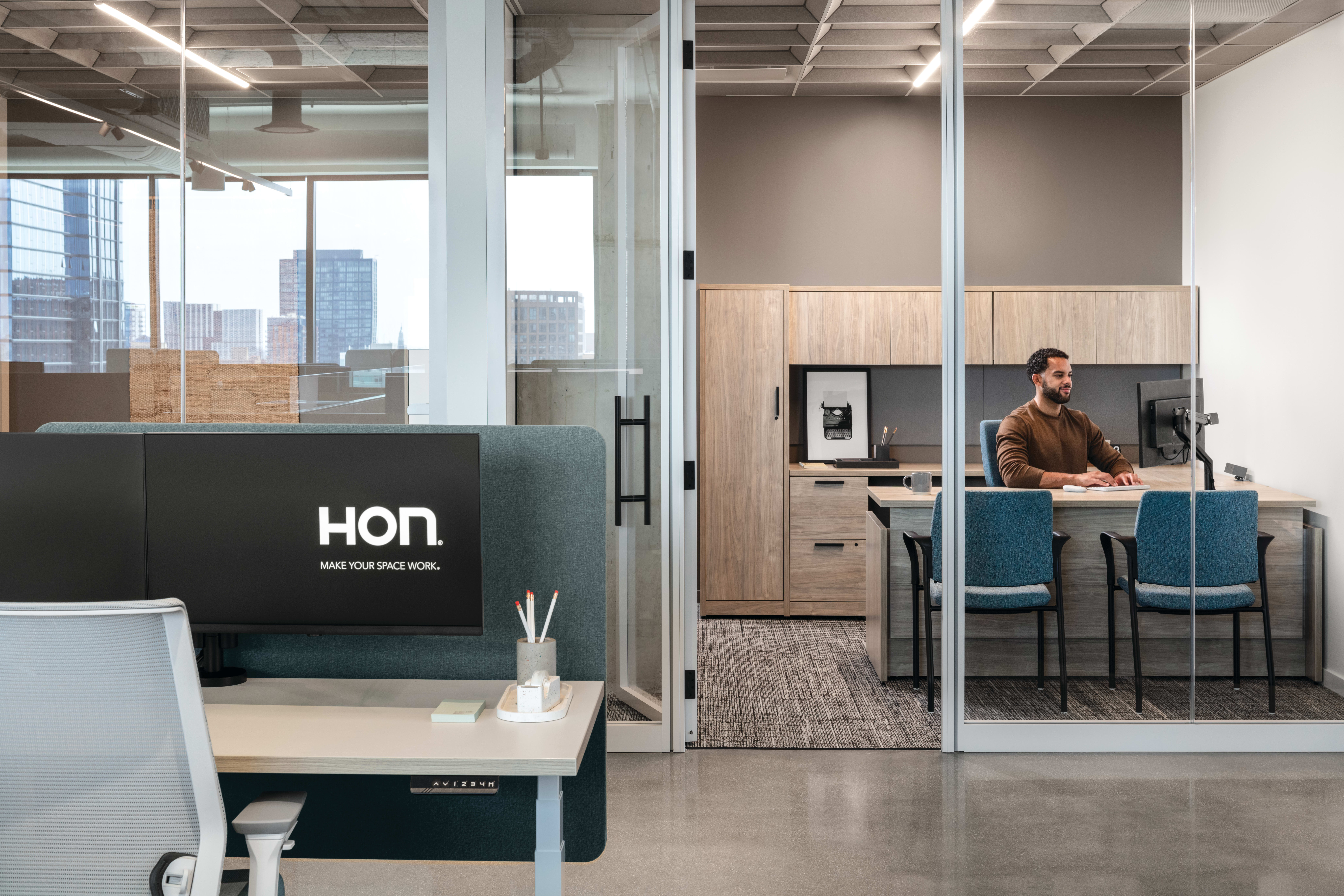 Open workstation with height-adjustable desk, privacy panel, task chair, and monitor, paired with glass-enclosed office featuring L‑shaped work surface, storage cabinets, and guest seating.