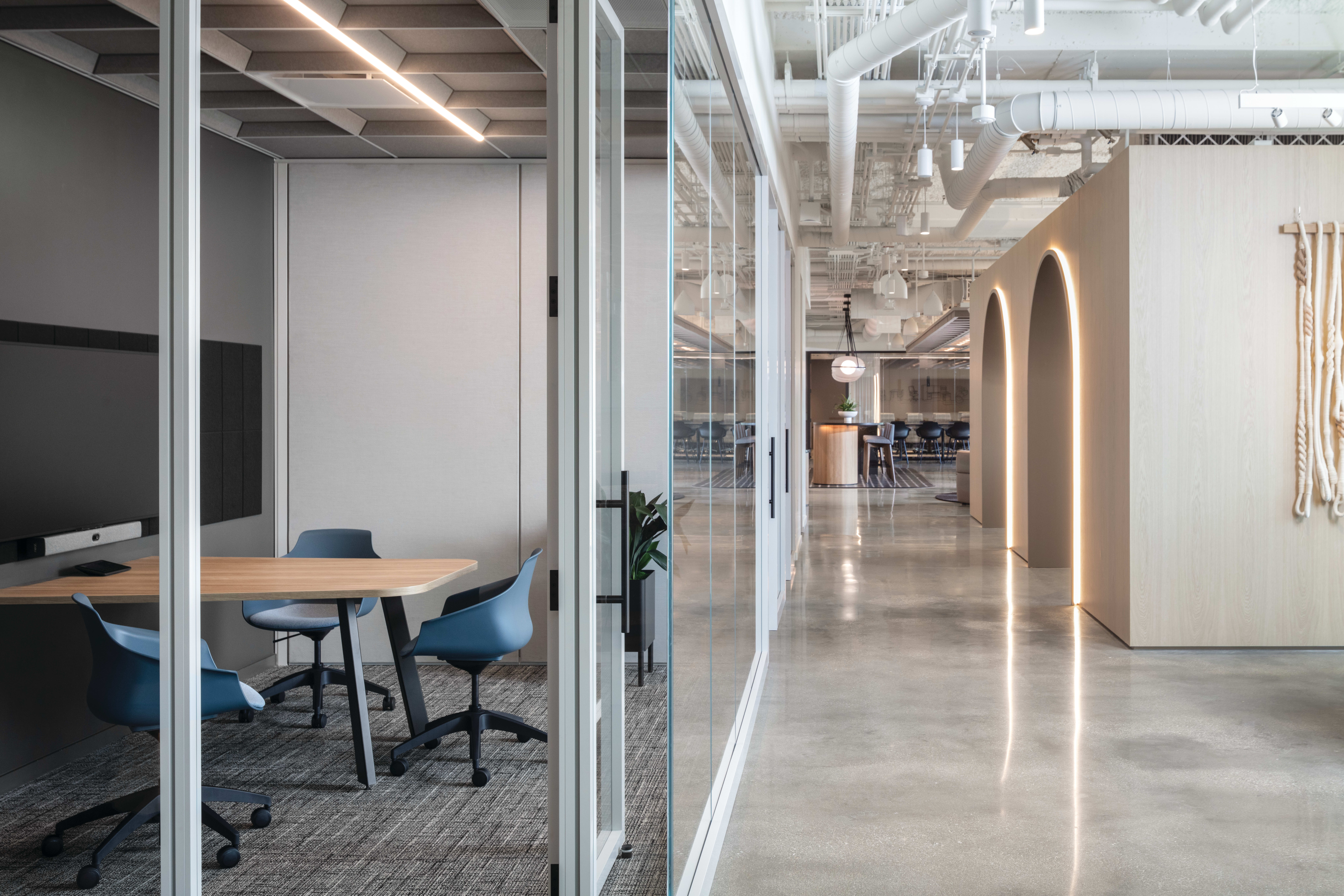 Glass-enclosed meeting room with round wood table, rolling task chairs, wall-mounted display, and adjacent lounge seating with additional tables and chairs.