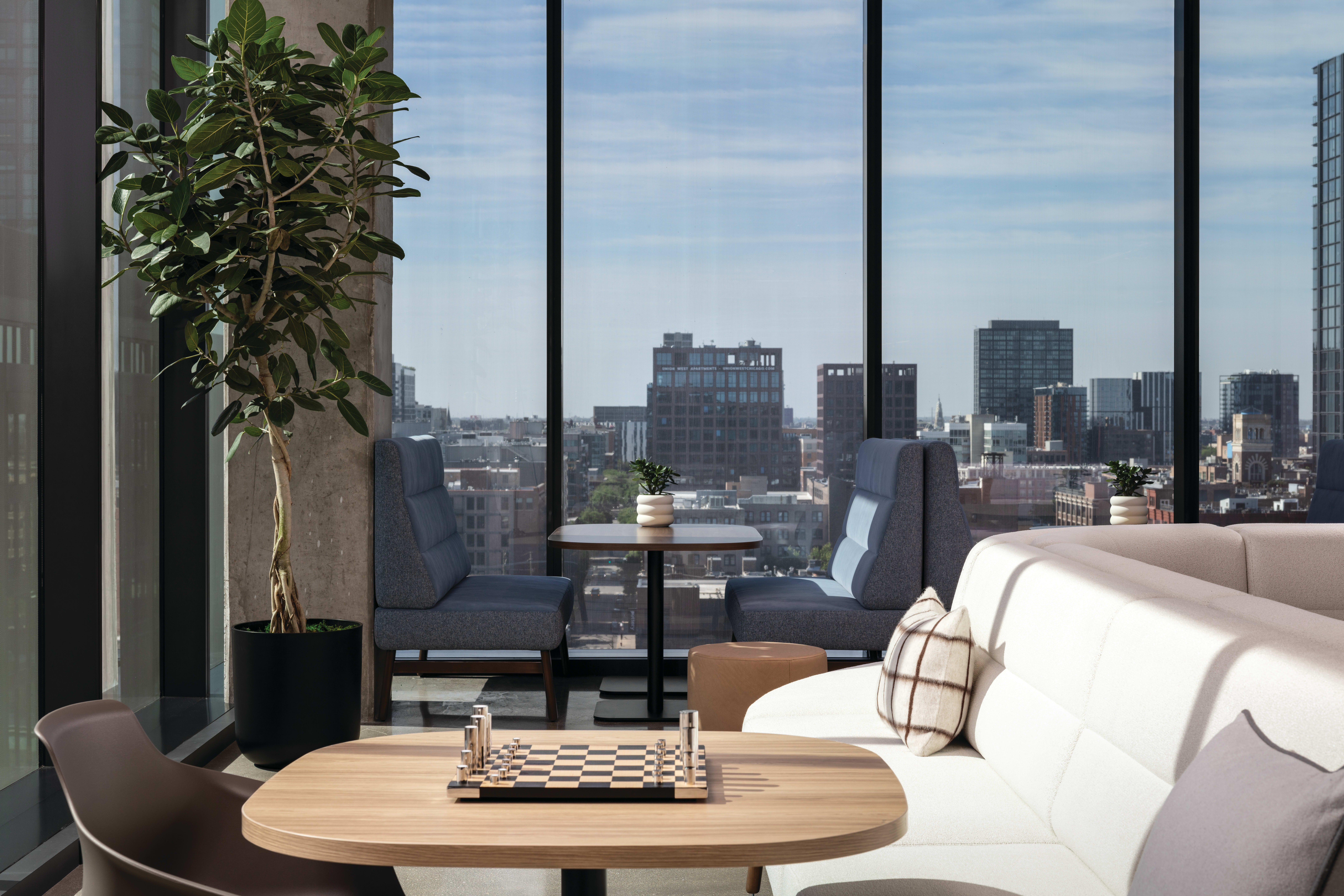 Modern lounge seating with small table, indoor plant, and floor-to-ceiling windows overlooking urban skyline.