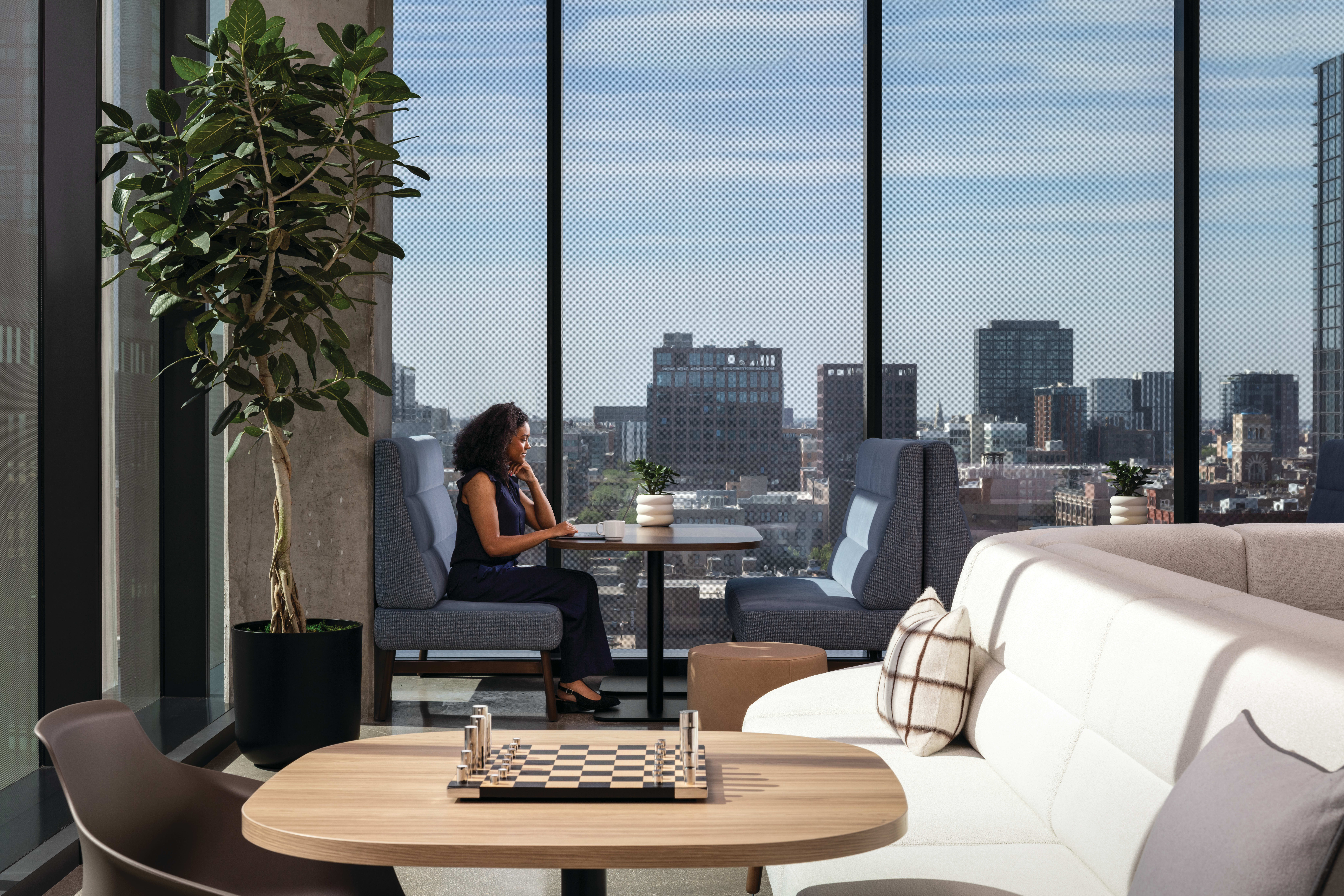 Modern lounge seating with small table, indoor plant, and floor-to-ceiling windows overlooking urban skyline.
