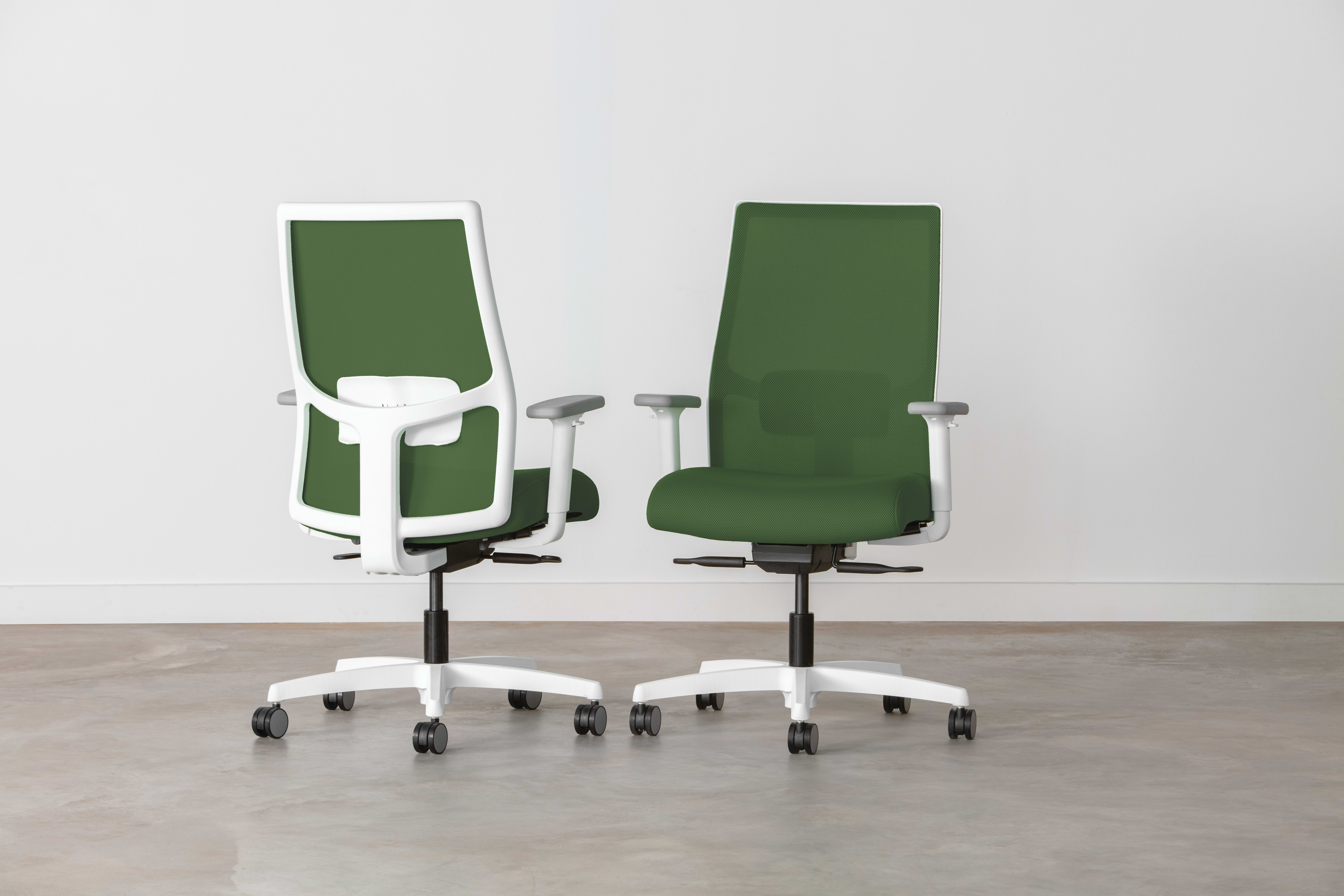 Two green and white office chairs with adjustable features are positioned in a minimalist, white-walled setting.