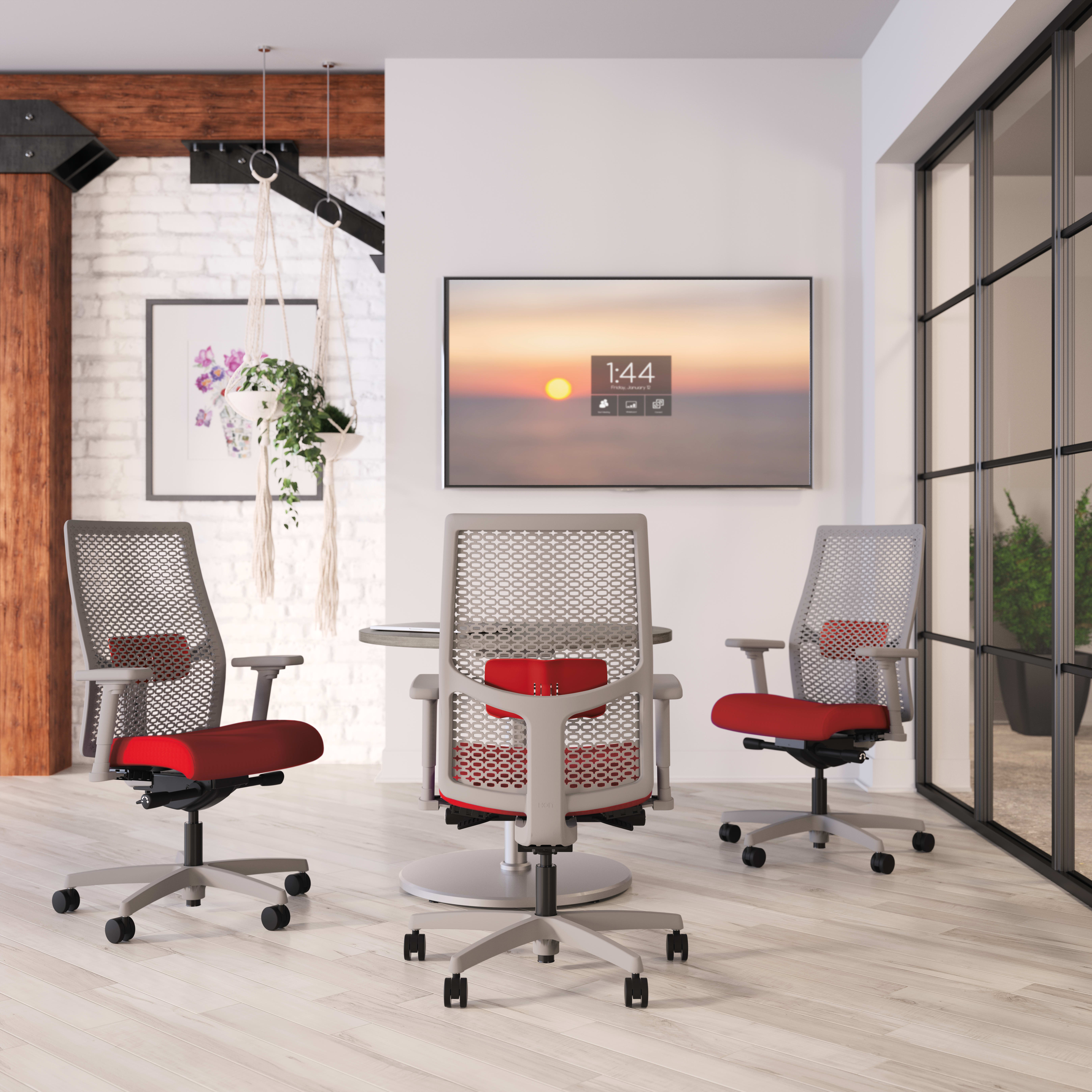 The image depicts a modern office space with white walls, wooden accents, and large windows overlooking a scenic sunset. In the foreground, there are several red and gray office chairs arranged around a table.
