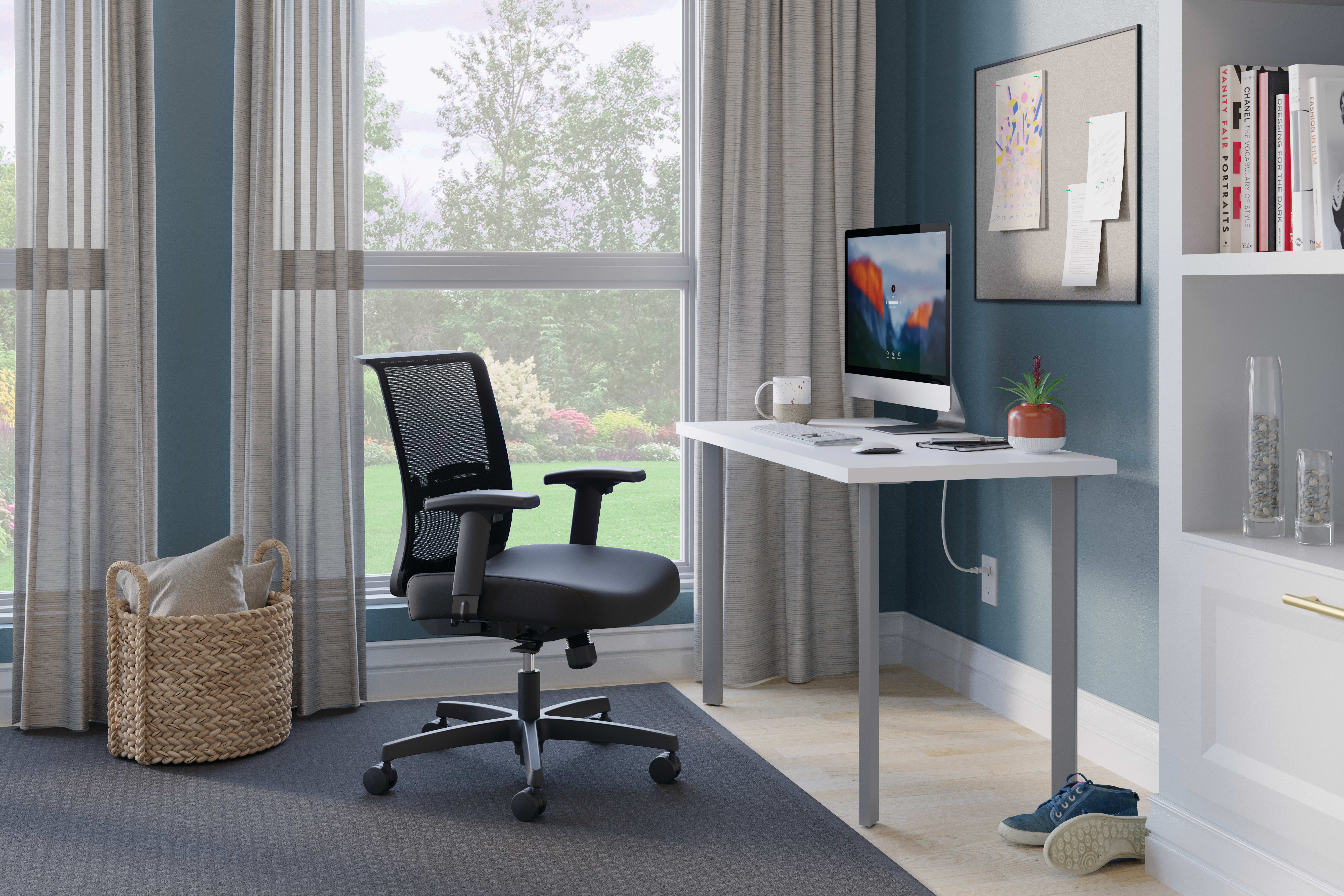 A modern home office with a white desk, a black ergonomic office chair, and various decorative elements such as artwork, plants, and a woven basket, set against a backdrop of large windows overlooking a lush outdoor scene.
