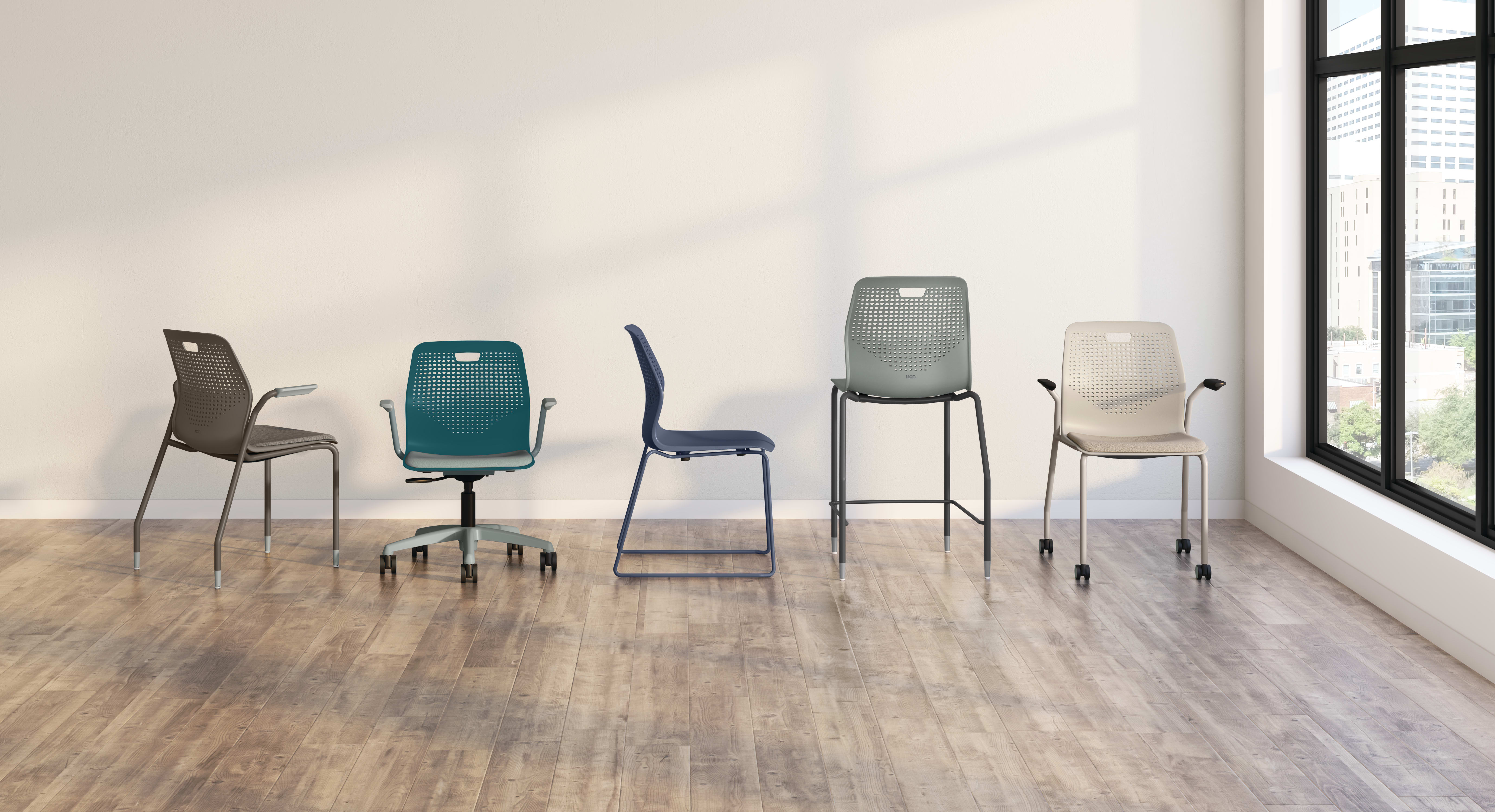 A variety of modern, minimalist chairs in different colors and designs are arranged in an empty, well-lit room with a wooden floor and a large window in the background.