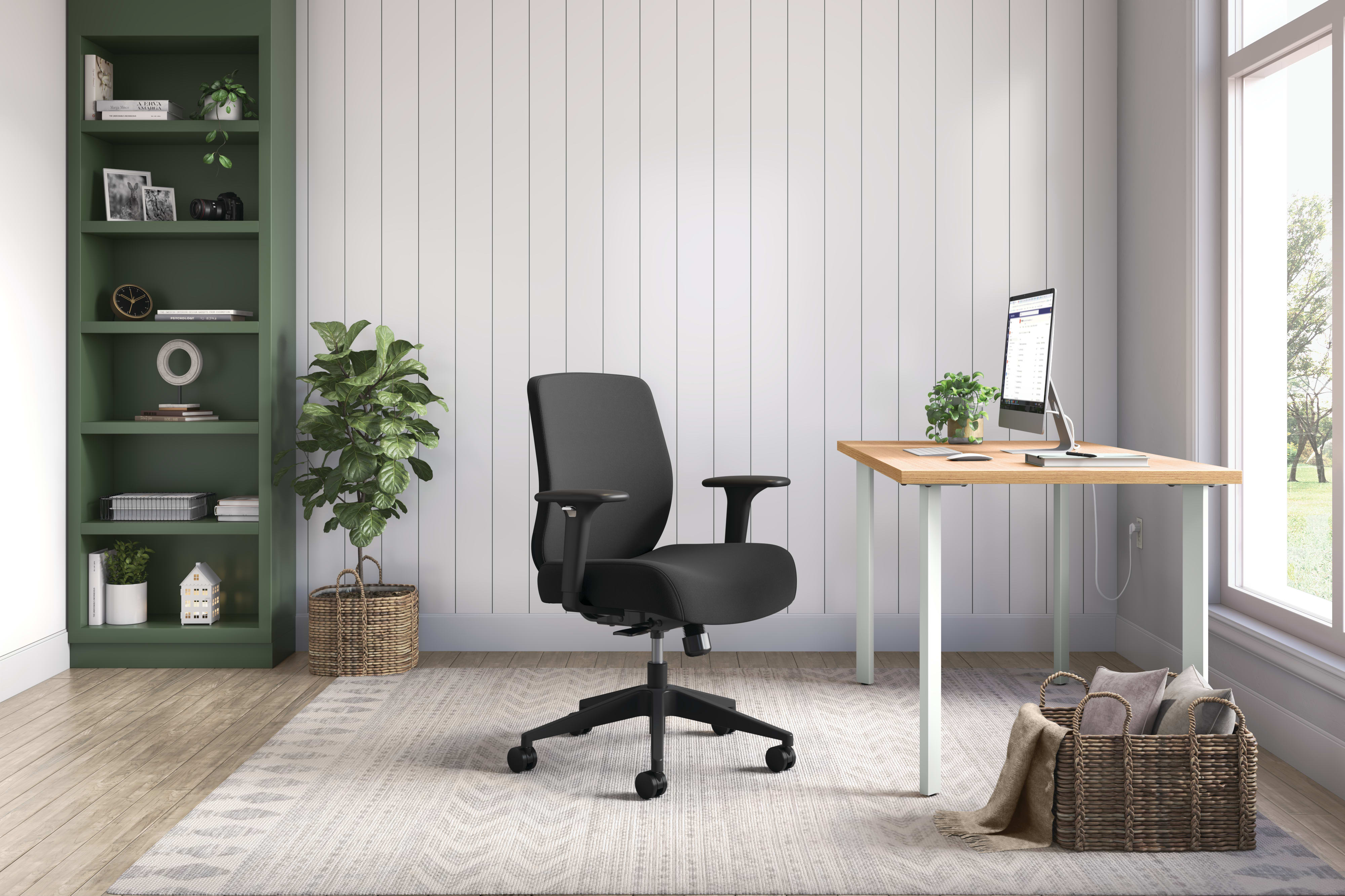 A modern and minimalist home office space with a wooden desk, a black office chair, and a green shelving unit against a white paneled wall.
