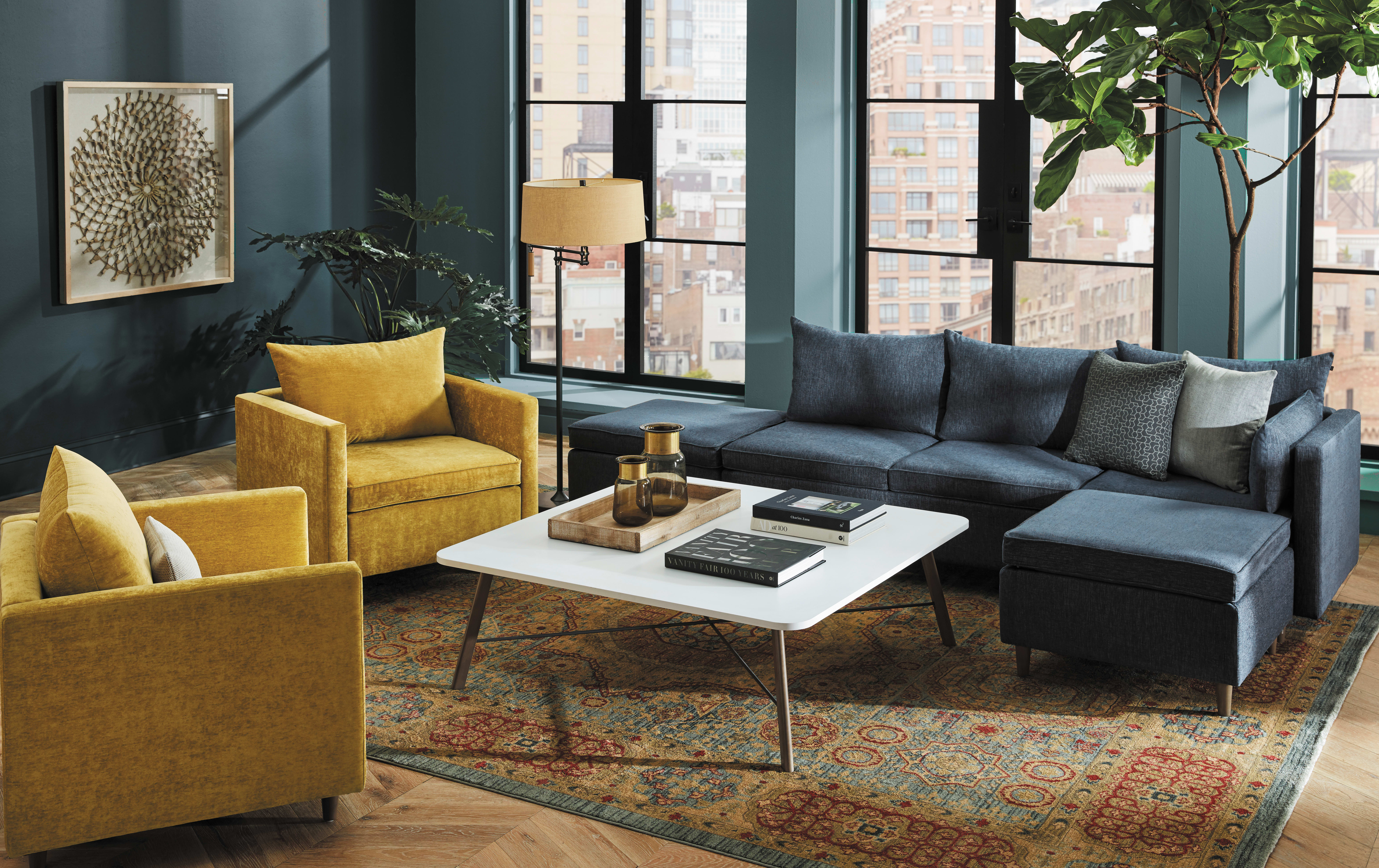 A cozy and stylish living room with a gray sectional sofa, yellow armchairs, a white coffee table, and a patterned area rug, set against a backdrop of large windows and lush greenery.
