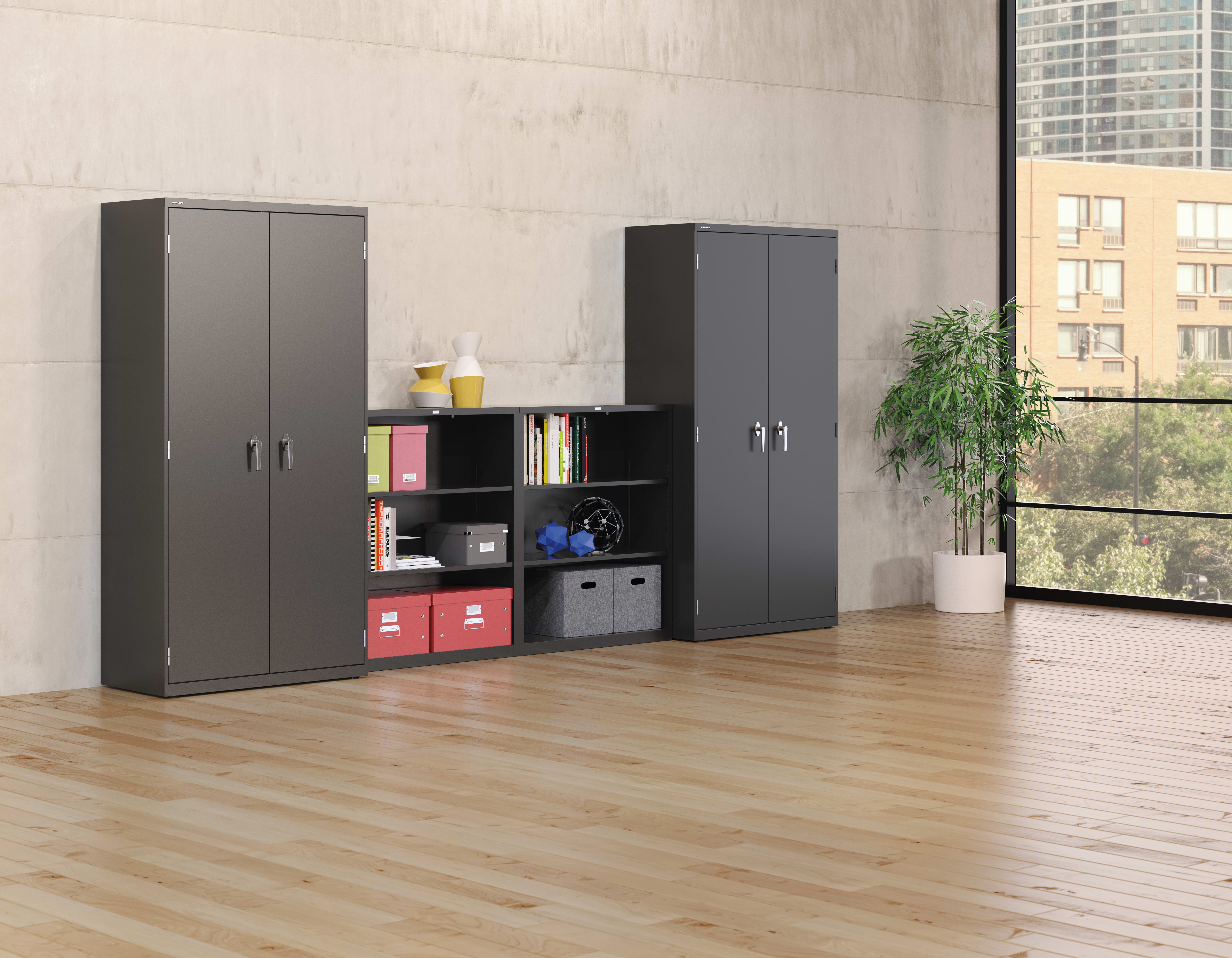 The image depicts a modern and minimalist office or home interior, featuring a set of black storage cabinets with shelves and compartments, arranged against a plain concrete wall. The floor is made of light-colored hardwood, and a potted plant adds a touc