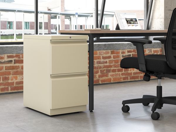 A modern office workspace with a wooden desk, a beige filing cabinet, and a black office chair set against a backdrop of exposed brick walls and large windows.