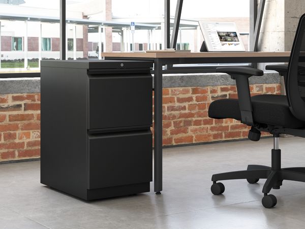 A modern office workspace with a black filing cabinet and a swivel chair in the foreground, set against a backdrop of brick walls and large windows.