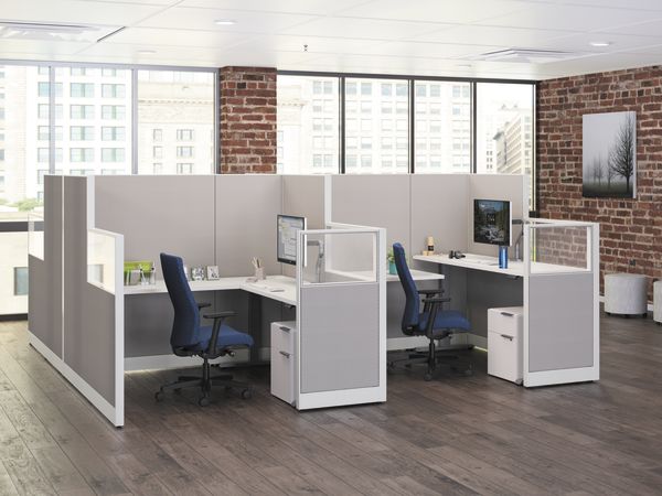 The image depicts a modern office space with cubicles, desks, and computers, set against a backdrop of large windows and exposed brick walls.