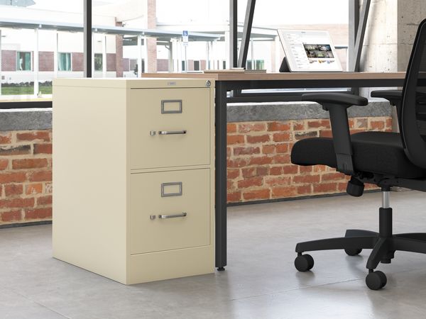 A beige filing cabinet stands in the foreground, with a desk and office chair visible in the background, set against a brick wall.