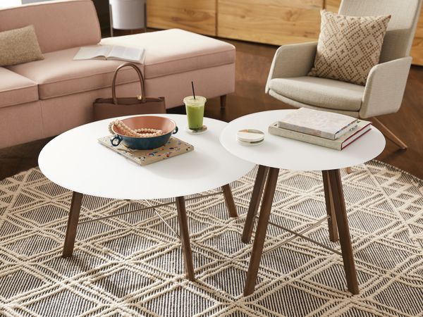 A cozy living room with a white round coffee table, a green smoothie, and a book on a patterned rug, surrounded by a beige sofa and a wooden cabinet in the background.
