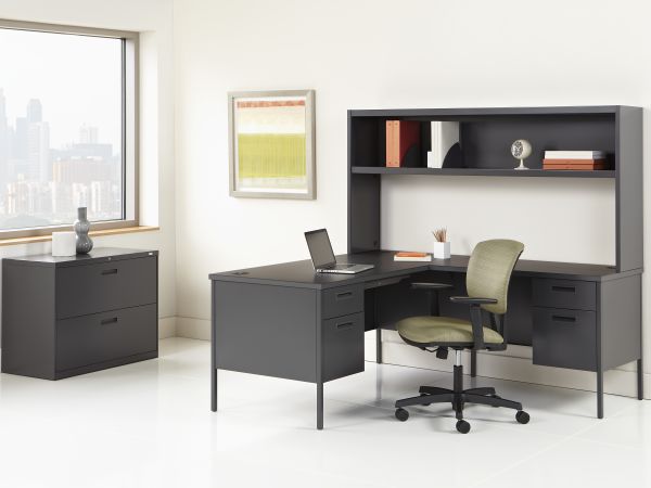 A modern and minimalist home office setup with a black desk, shelving unit, and a green office chair against a white wall with a large window.