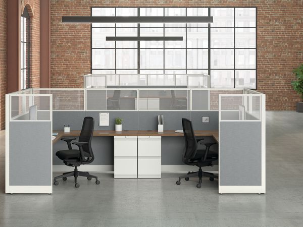 A modern, minimalist office space with white and gray cubicles, chairs, and a potted plant against a brick wall background.