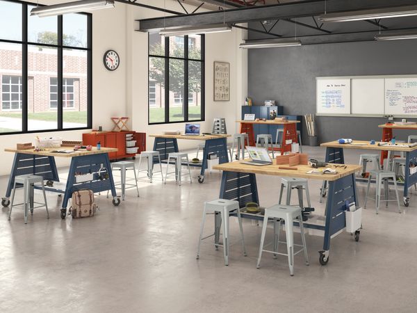 A modern, open-concept workspace with various workstations, desks, and chairs arranged throughout the room. The space features large windows, exposed ceiling beams, and a whiteboard on the wall, creating a bright and collaborative environment.