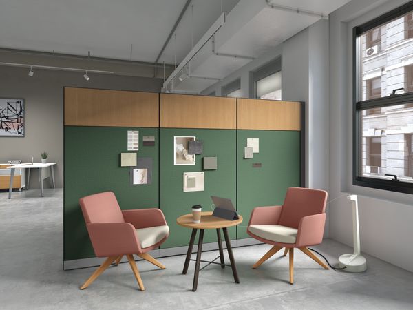 Office seating arrangement with two pink armchairs, round wooden table, and green partition wall with notes.