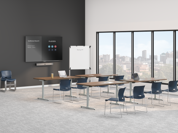 A modern training room with long tables, blue chairs, a wall-mounted display screen, a whiteboard, and large windows overlooking a city skyline.