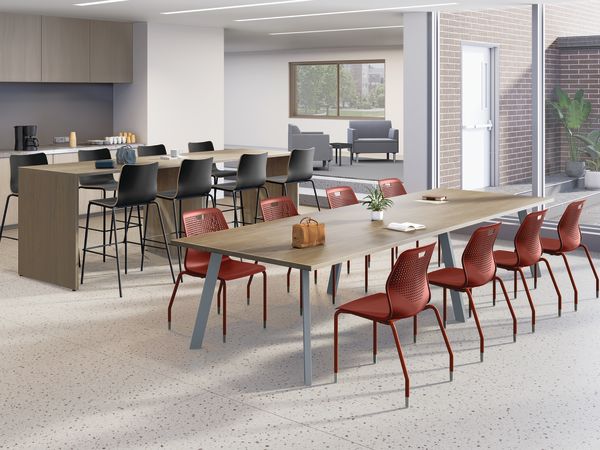 Modern collaborative workspace with long wooden tables, red and black chairs, coffee station, adjacent lounge seating, large windows, and bright contemporary lighting.