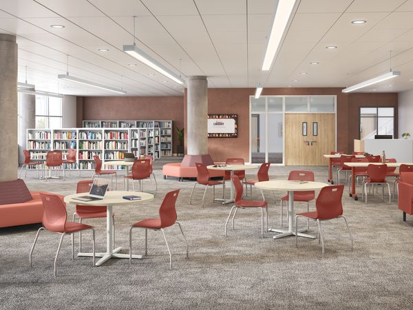Modern library commons with bookshelves, round study tables, red chairs, lounge seating, structural columns, carpeted flooring, and bright overhead lighting in an open interior space.
