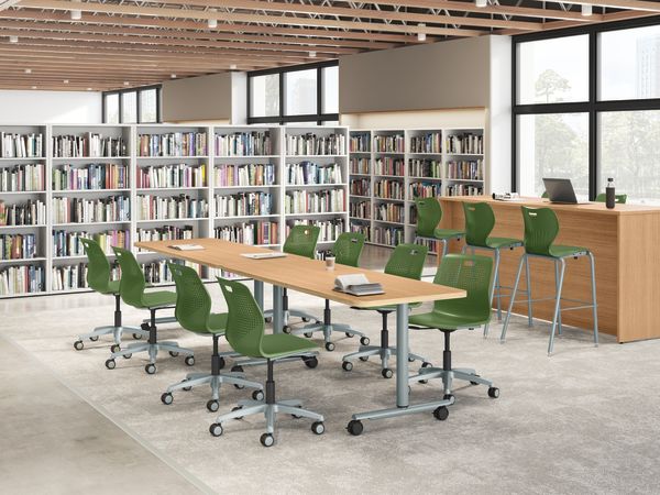 Modern library study area with tall bookshelves, large communal tables, green task chairs on casters, high counter seating, expansive windows, wood ceiling slats, and bright natural light.